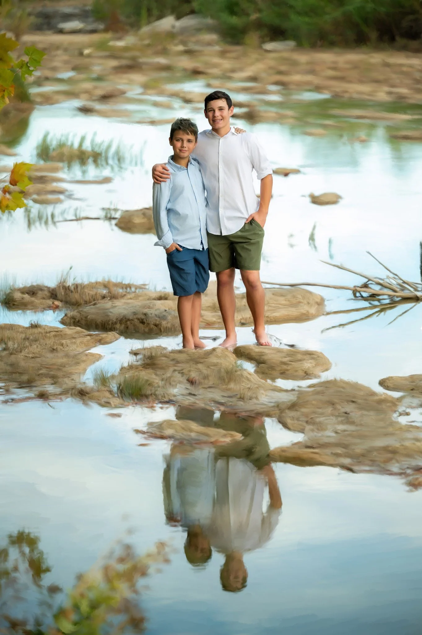 Two boys standing on rocks in a creek, smiling, with their reflection visible in the water.