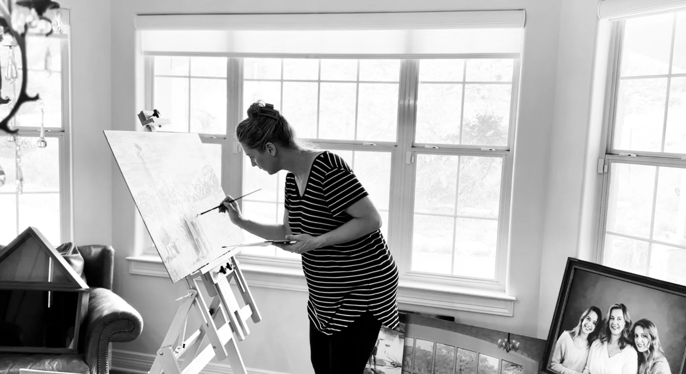 A woman in a striped shirt painting on a canvas in a bright room with large windows, with artwork and a framed photograph of three women nearby.