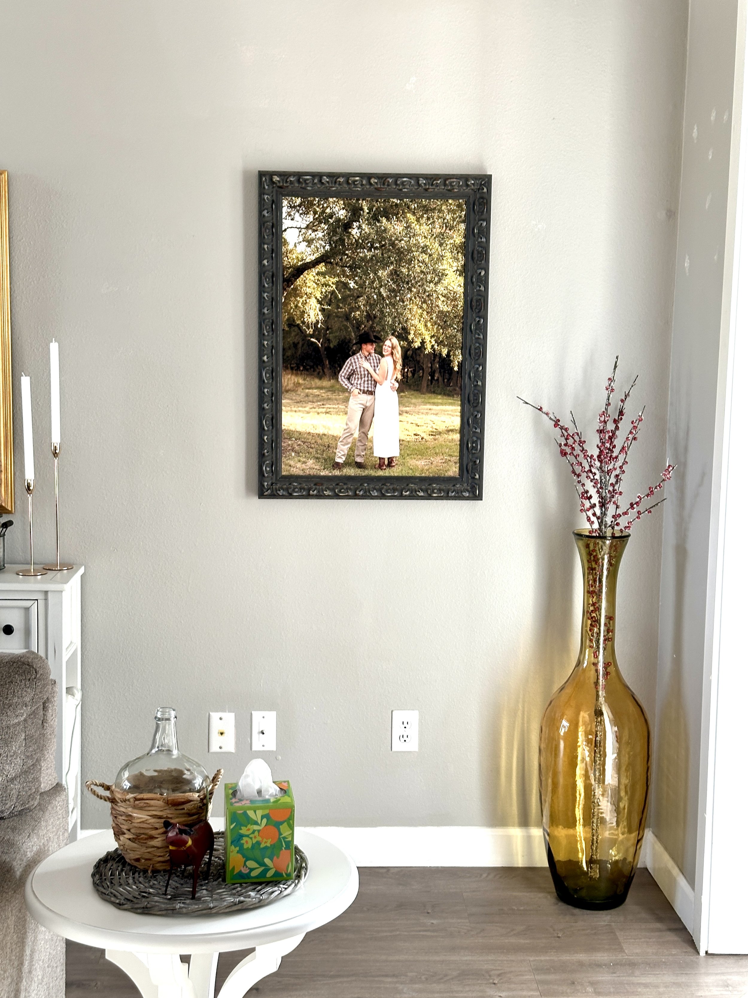A living room wall with a framed photo of a couple outdoors in a park, a tall yellow glass vase with red branches, a small white side table with a woven basket, a glass jar, a decorative figure, and a tissue box.