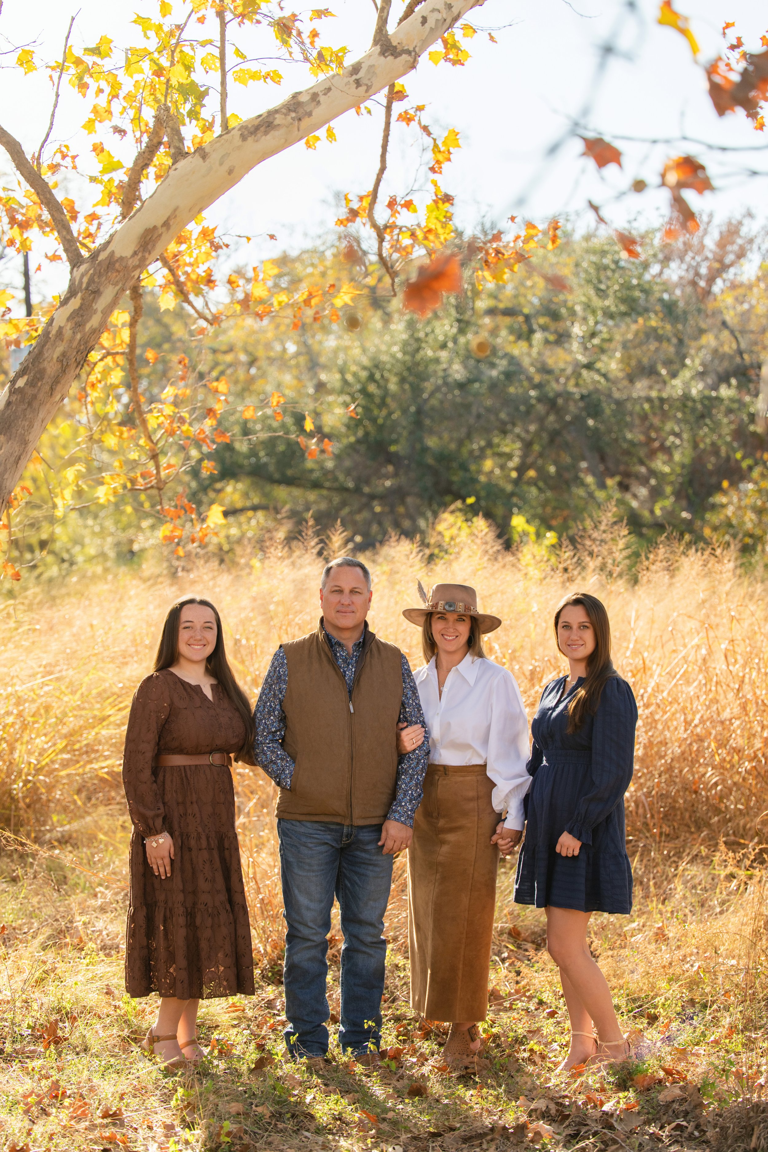 A family portrait with the parents and two grown daughters standing in a golden field with gold and orange oak leaves in the background, by Wimberley photographer Heidi Knight.