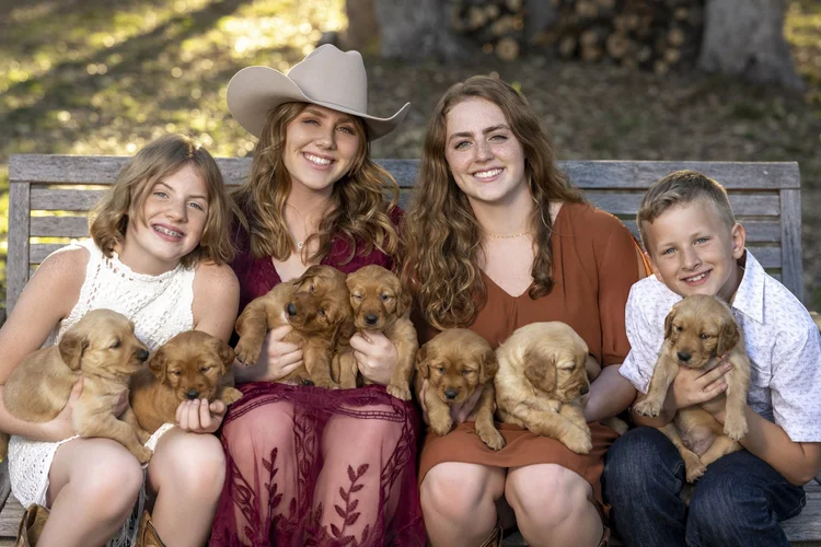 Four children and two women sitting on a wooden park bench outdoors, each holding a golden retriever puppy.