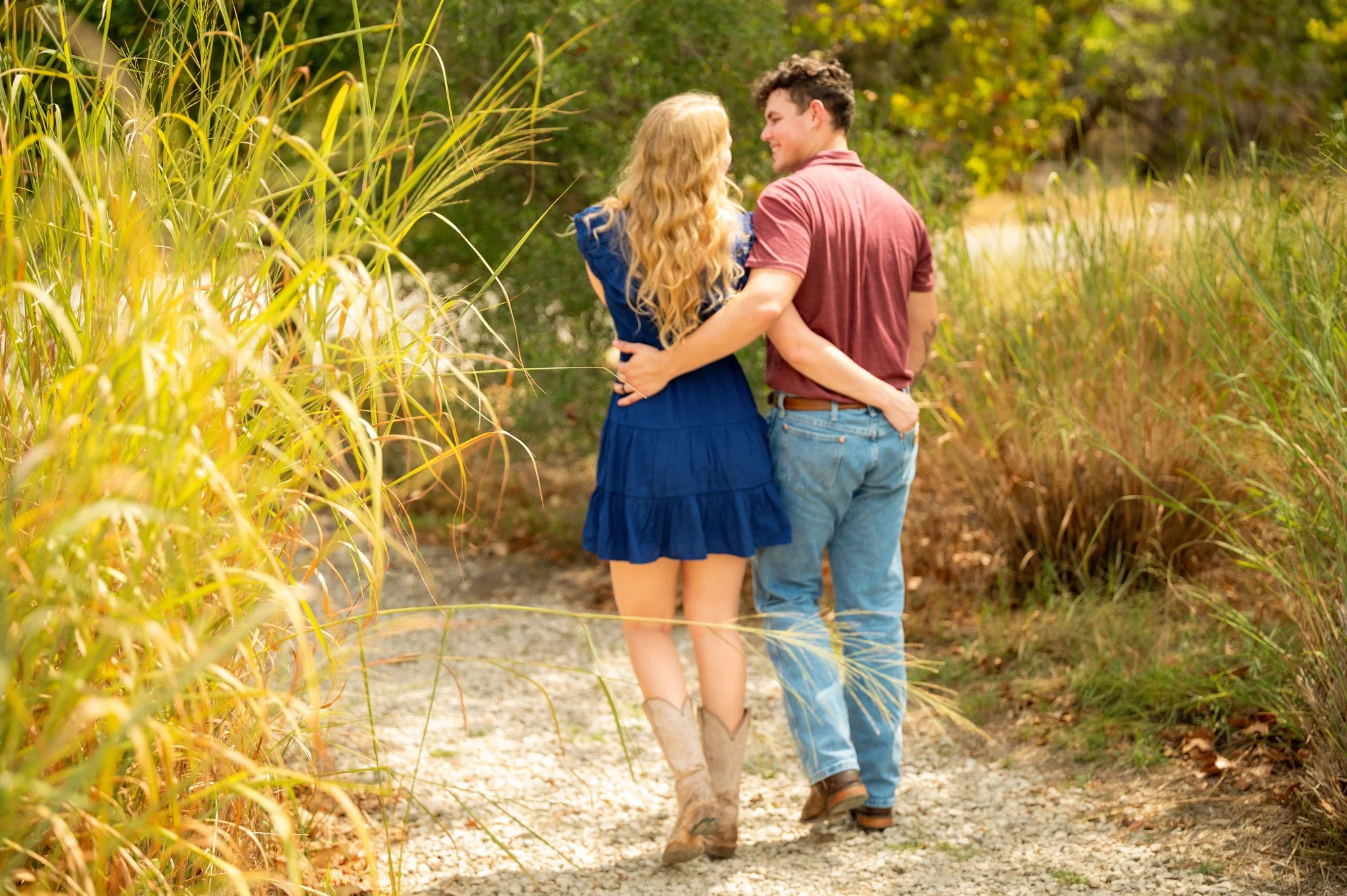 A young couple walking on a dirt path through tall grass and trees, embracing and smiling at each other.