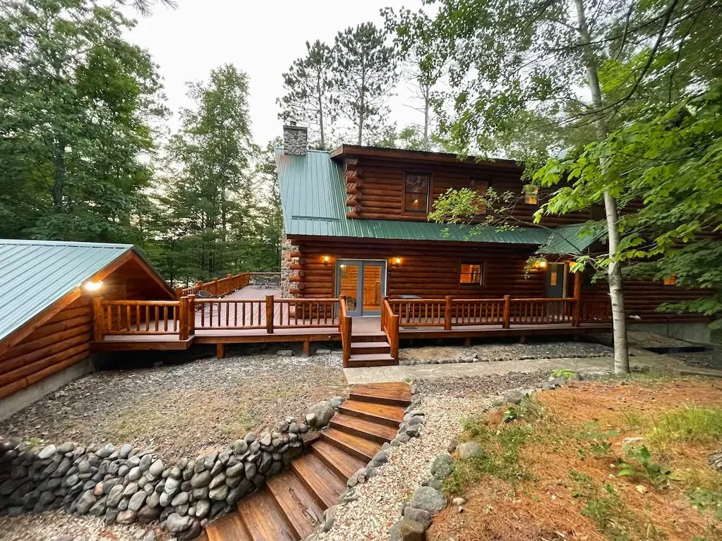Wooden house with a green metal roof surrounded by trees, with an elevated wooden deck and stone steps leading up to it.