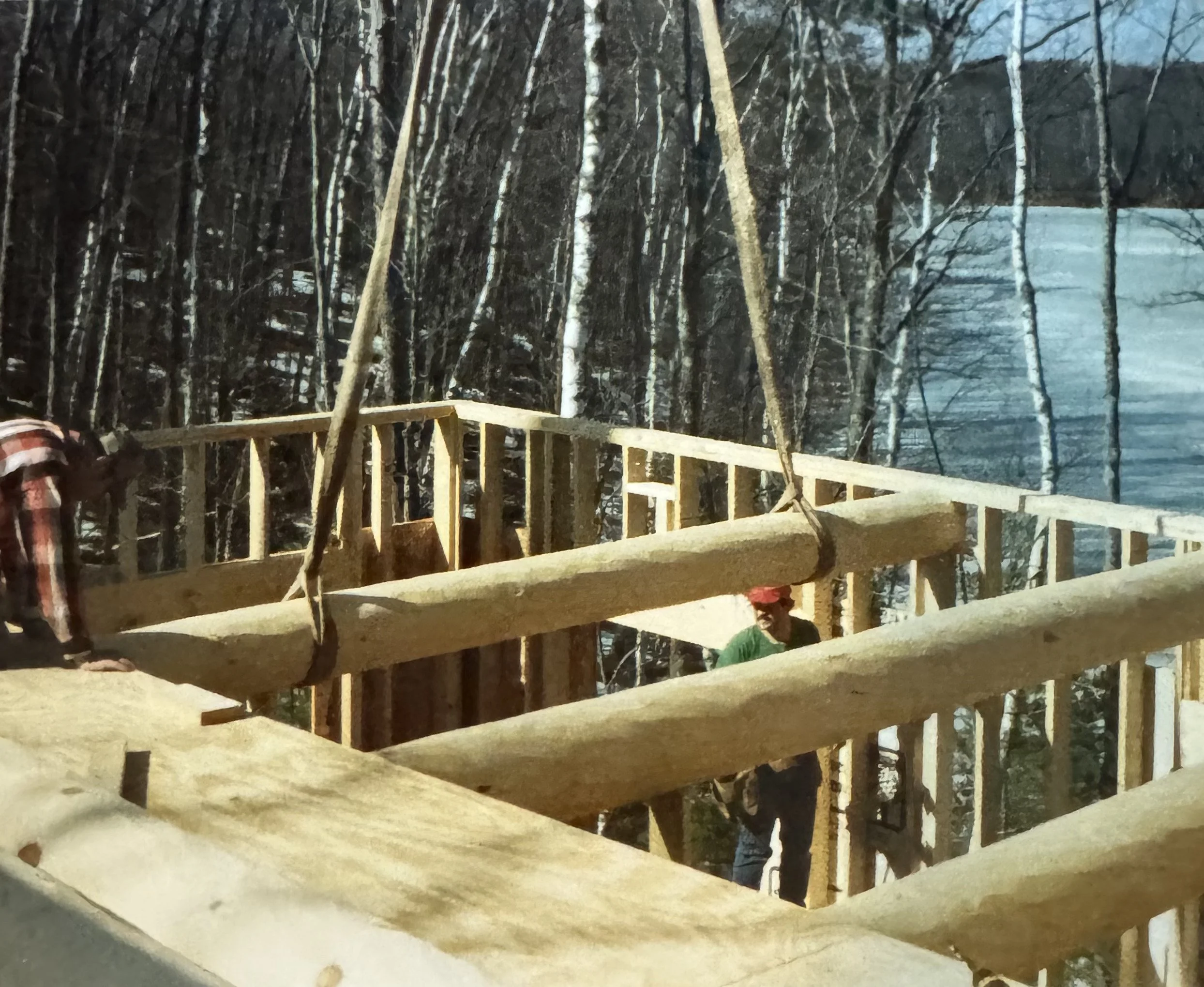 Wooden frame of a building under construction in a forested area with some workers visible, trees in the background, and a body of water nearby.