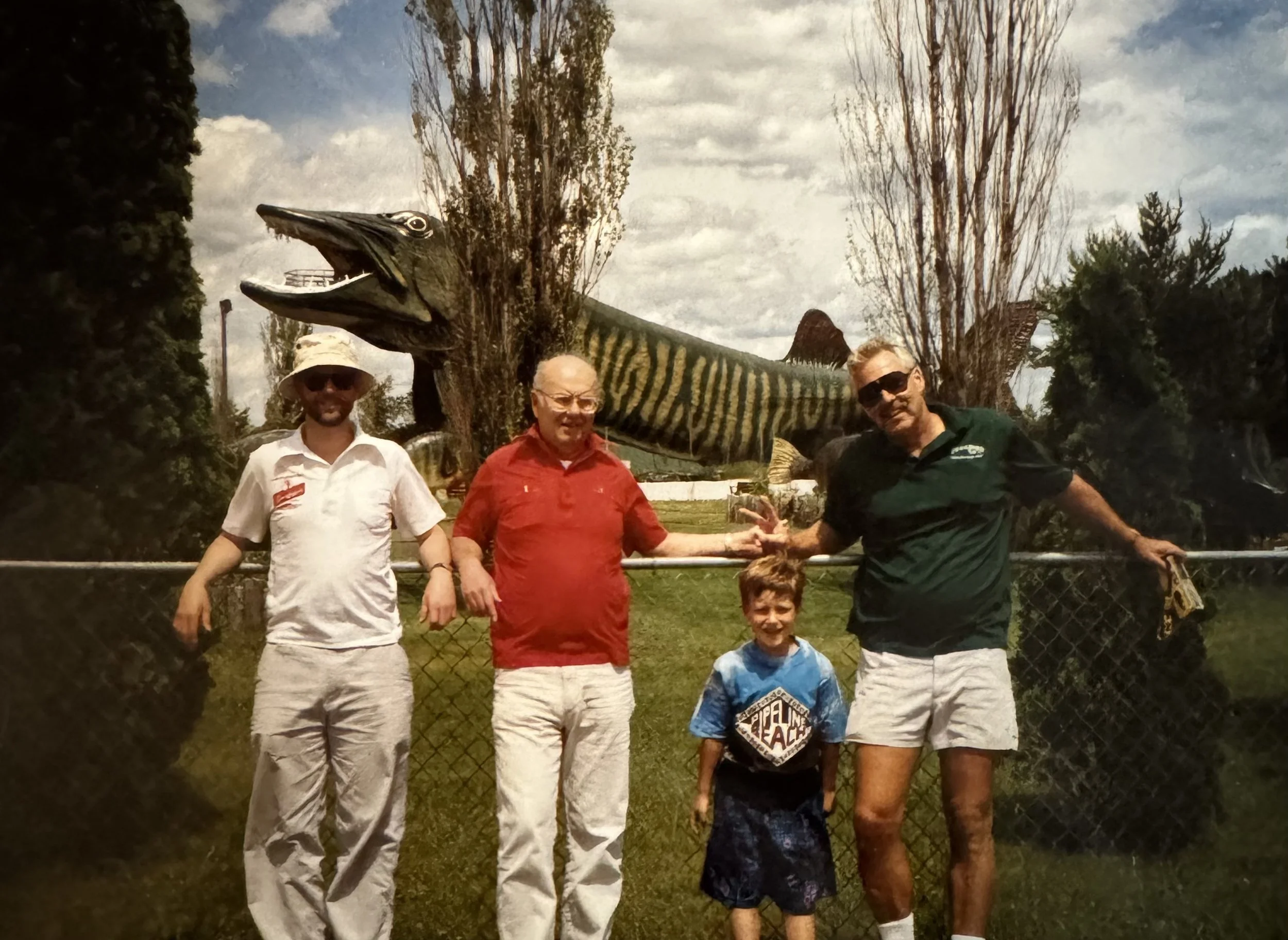 Four people standing in front of a chain-link fence, with a large dinosaur sculpture behind them. The group includes two men, a woman, and a young boy, all smiling and holding hands. The dinosaur appears to be a large, predatory species with an open mouth, displayed outdoors on a cloudy day with trees in the background.
