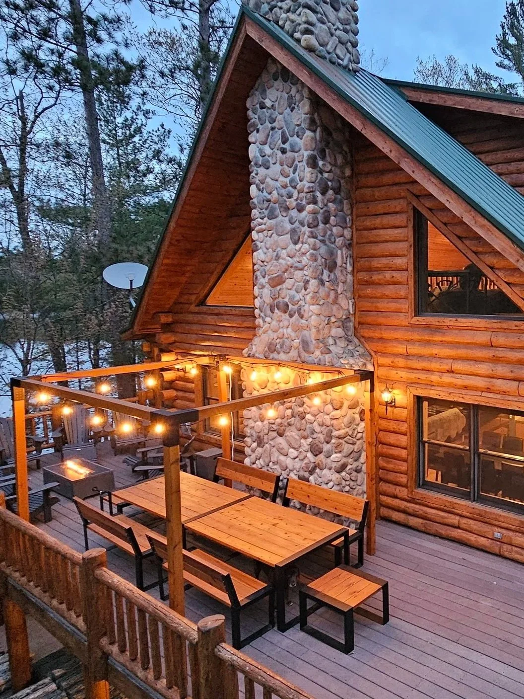 A wooden cabin with a large stone chimney and a deck decorated with string lights, outdoor furniture, and surrounded by trees.