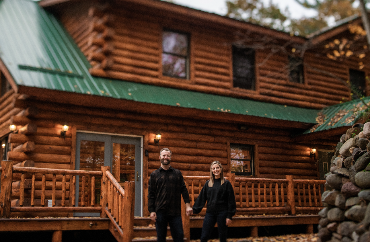 A smiling man and woman holding hands in front of a two-story log cabin with a green metal roof, wooden deck, and autumn foliage.