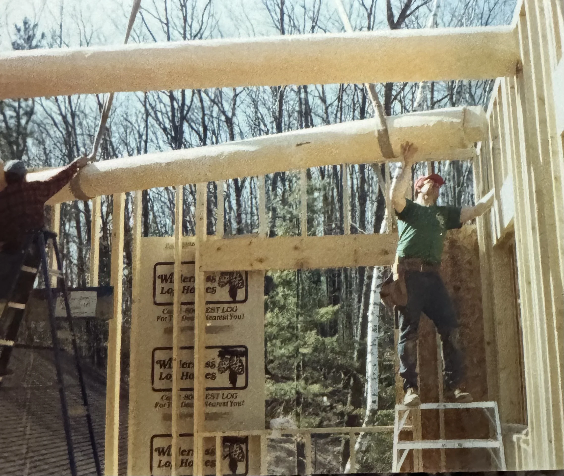 Two construction workers building a wooden structure outdoors, with trees in the background. One worker is on a ladder, and the other is standing on a platform, attaching wooden beams.