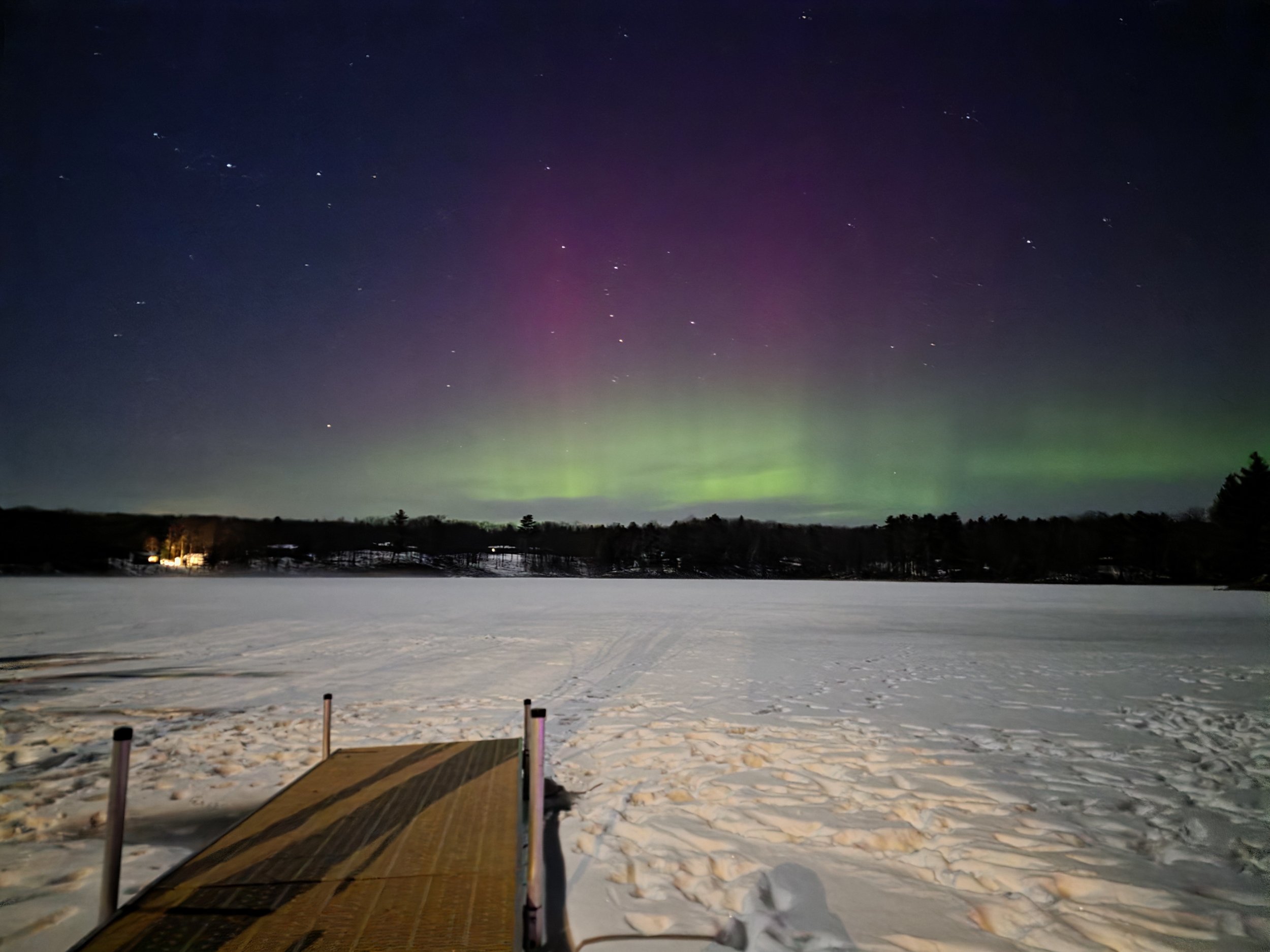 Nighttime view of the Northern Lights over a snow-covered landscape with a dock in the foreground and trees in the distance.