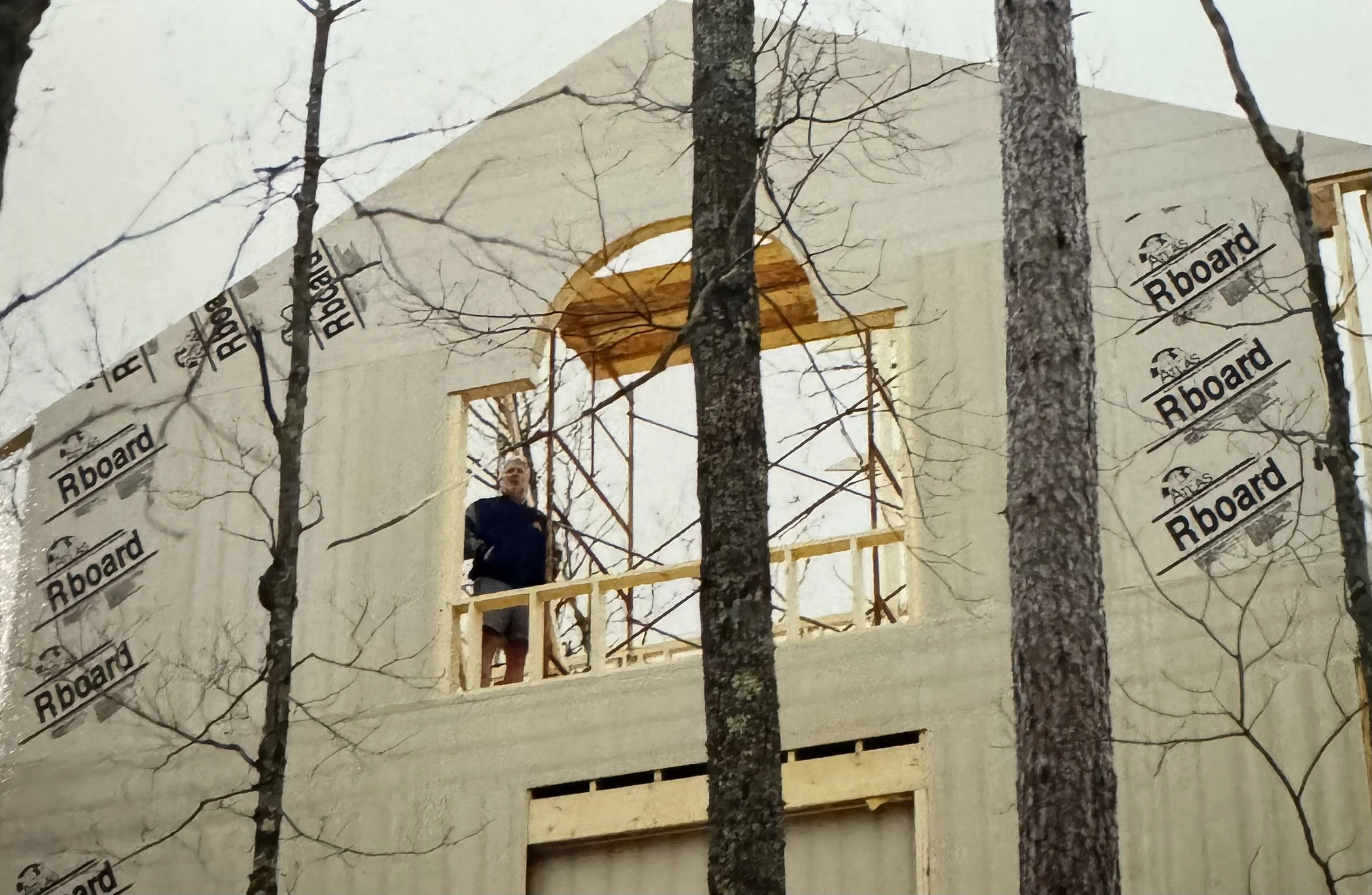A person standing on a construction balcony of a building in the process of being built, with trees in the foreground and a partially constructed facade with Rboard sheathing.