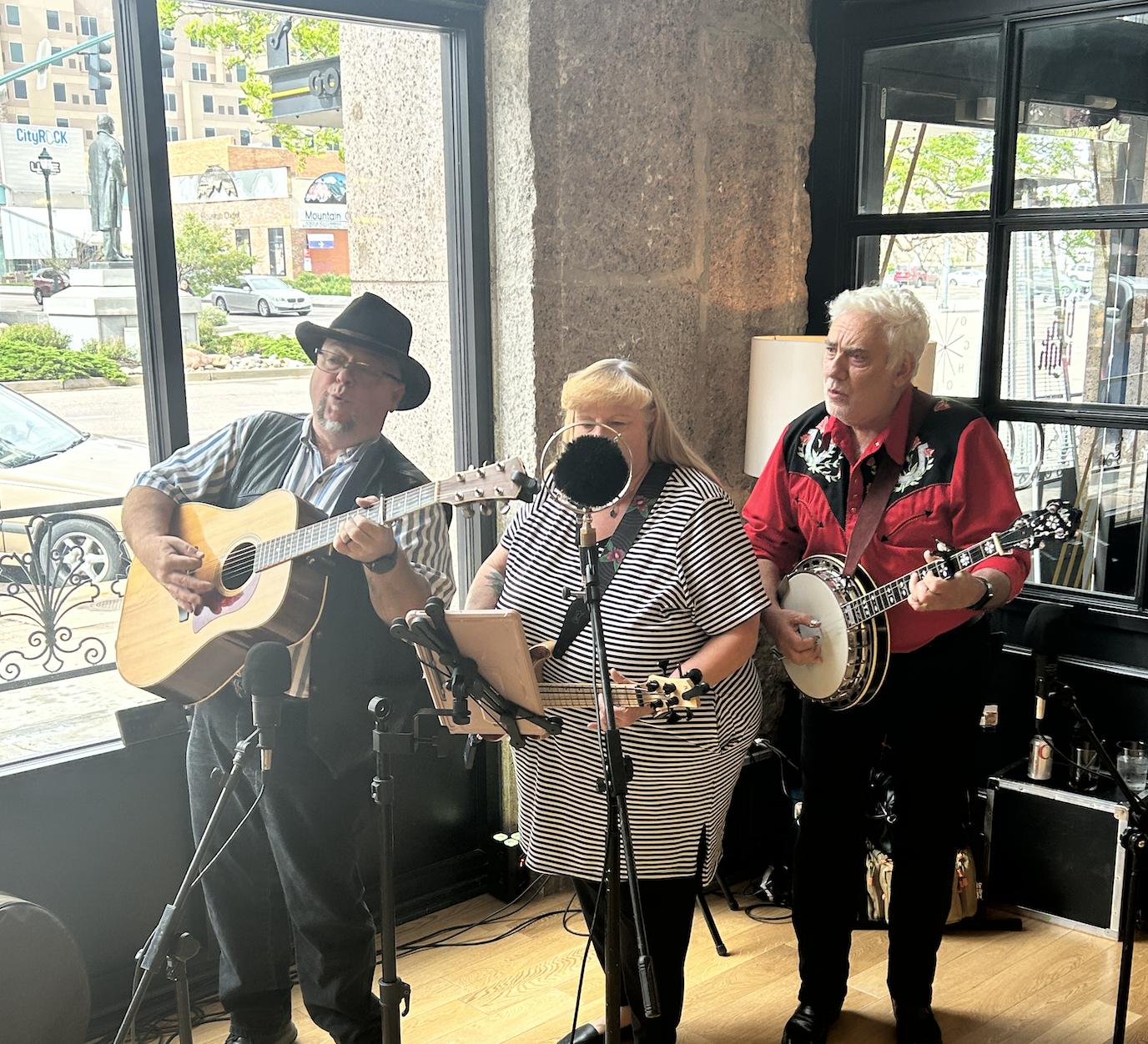 Three musicians performing indoors with a large window behind them. One man is playing a guitar, a woman is singing into a microphone, and another man is playing a banjo.