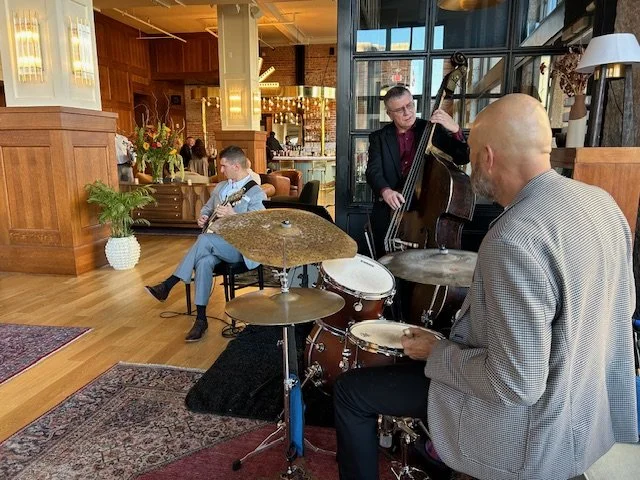 Three musicians playing in a cozy, warmly lit restaurant or lounge. A drummer with a light gray blazer, a standing double bassist in a dark suit, and a seated guitarist in a light blue suit. The seating area has wooden decor and large windows.