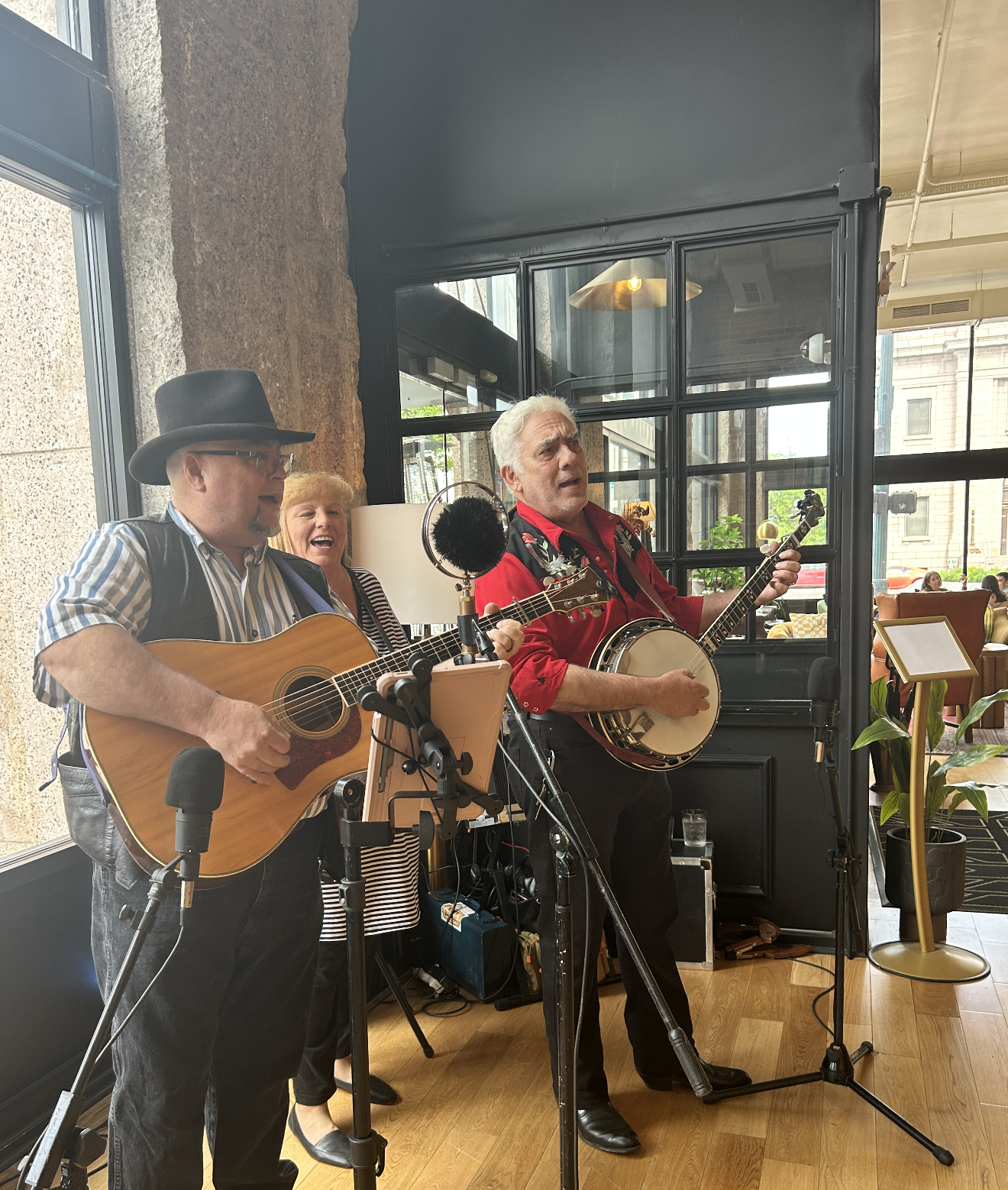 Three musicians performing with guitars and a banjo inside a restaurant, with large windows and modern decor.