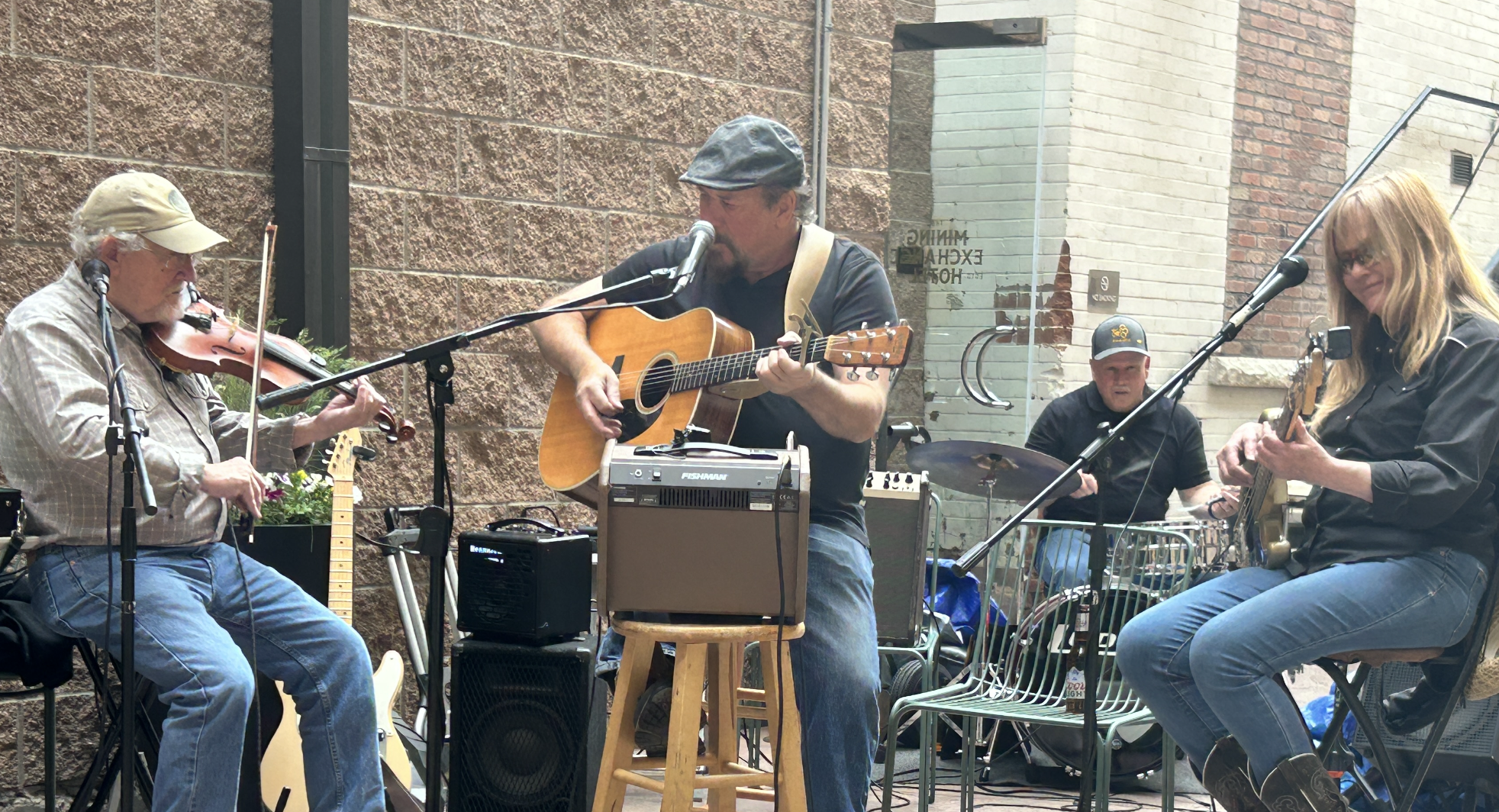 A group of four musicians playing instruments outdoors. One person is playing the violin, another is playing the acoustic guitar, a third is on drums, and the fourth is playing an electric guitar. They are performing against a brick wall and a storefront window.