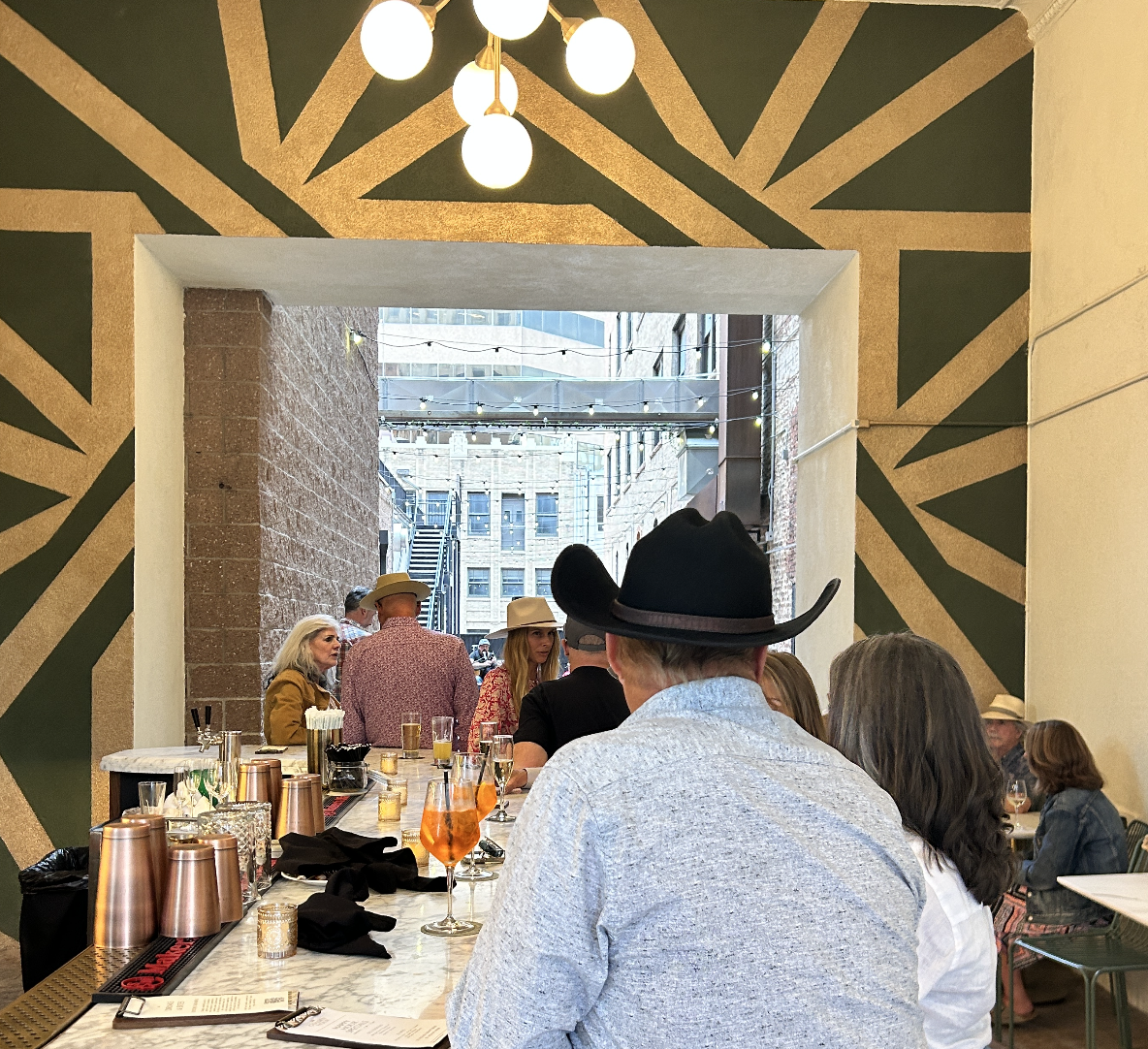 People dining at a bar with a decorative black and gold geometric pattern on the wall behind them, and a large opening leading to an outdoor area with brick and glass buildings.