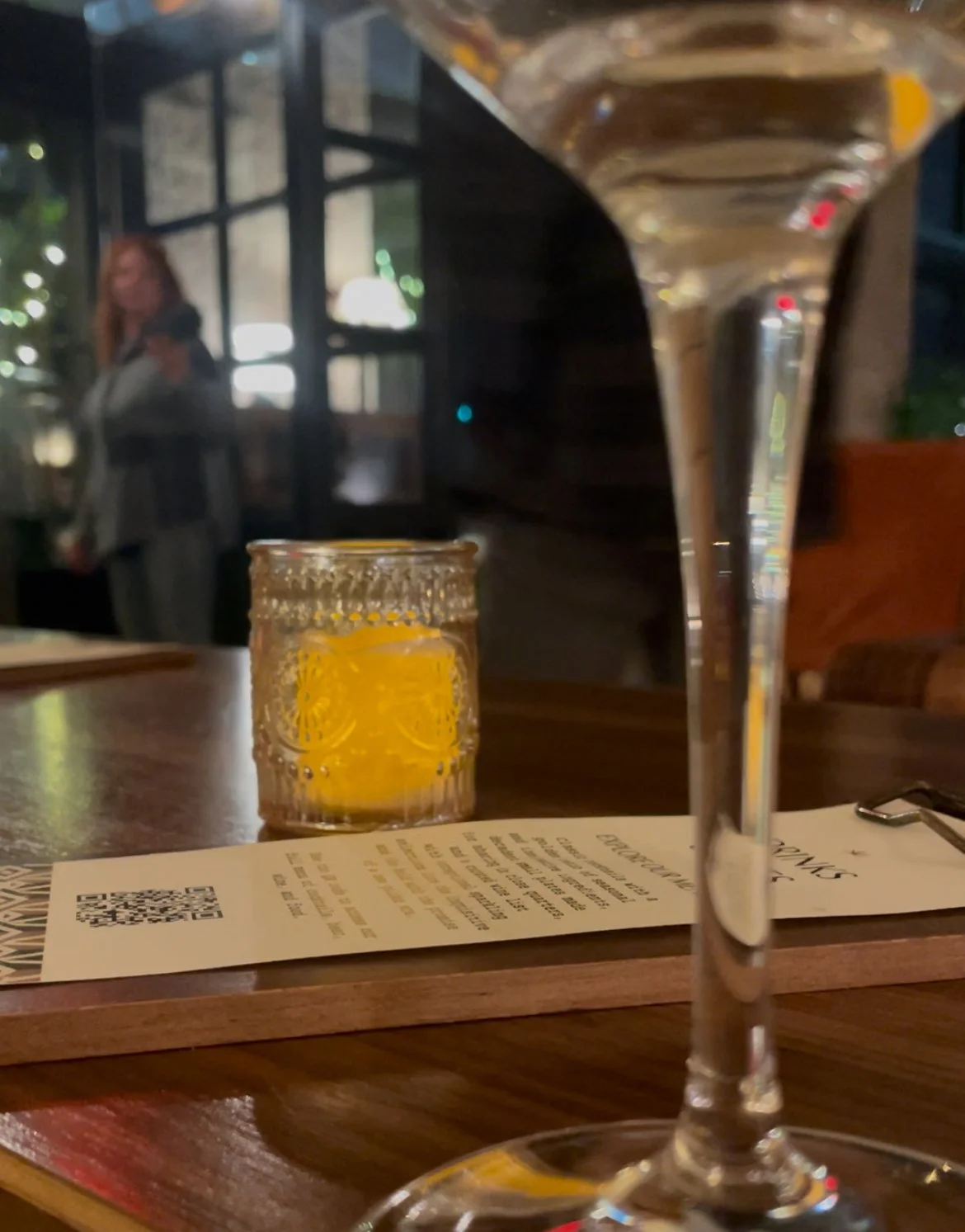 A dimly lit restaurant table with a large wine glass in the foreground, a small glass with lemon slices, and a paper menu on the tabletop. A blurry person stands in the background inside the restaurant.