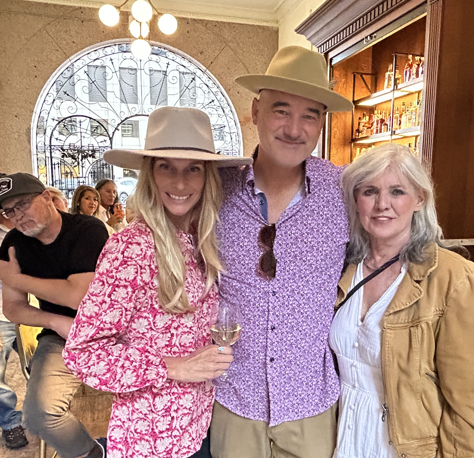The woman on the left is wearing a pink patterned blouse and a beige hat, holding a glass of white wine. The man in the middle is wearing a purple floral shirt, a beige hat, and sunglasses hanging on his shirt. The woman on the right has gray hair, a white shirt, and a tan jacket.