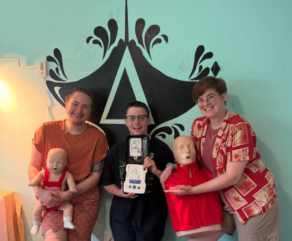 Casper and Cecíl pose with CPR mannikins as a student holds up an AED trainer. The student has successfully completed their Basic Life Support course!