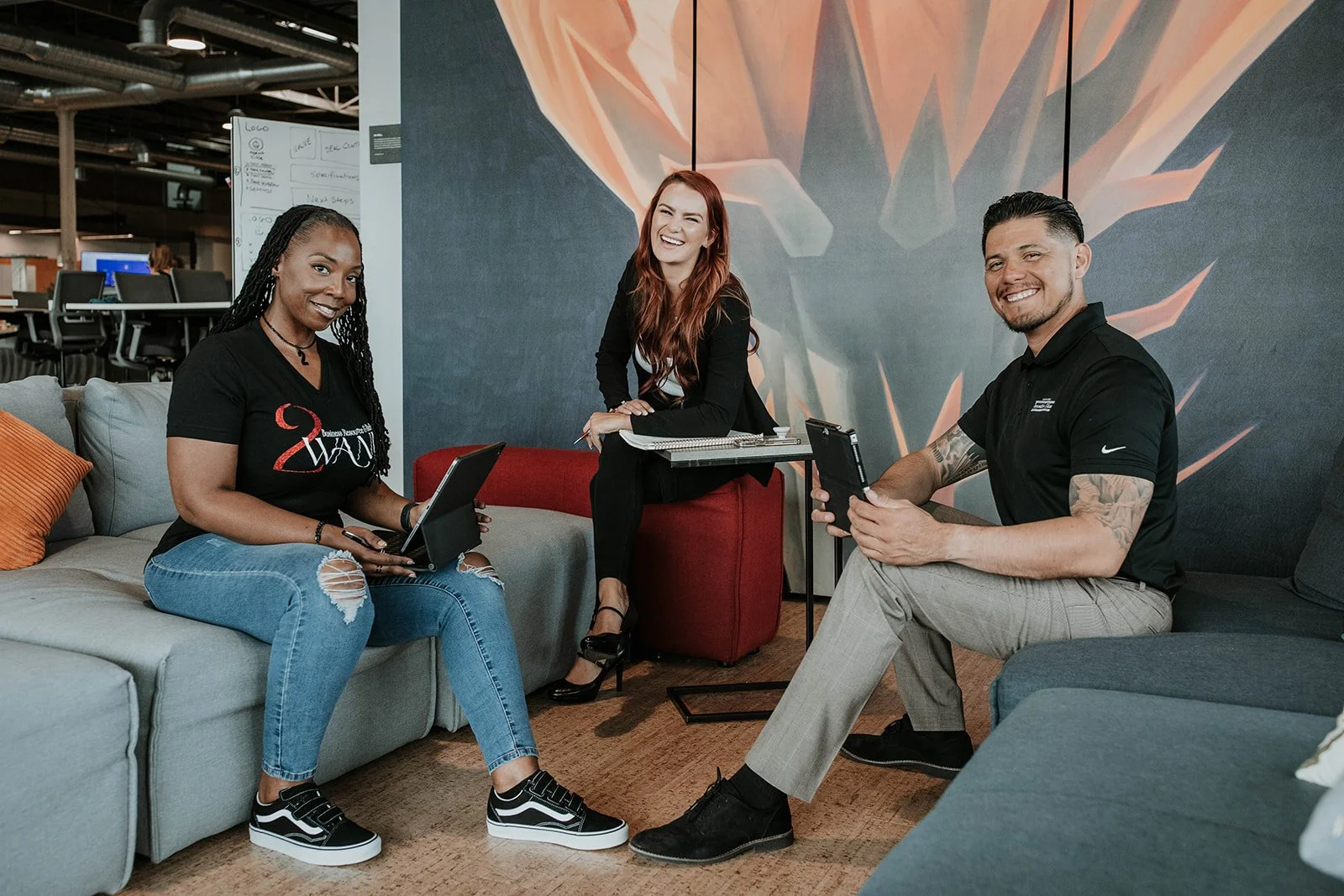 Three people, two women and one man, are sitting on couches in an office lounge area, smiling and holding tablets. A woman with long red hair is sitting on a red ottoman, and a woman with braided hair and a man with tattoos are sitting on the couches.