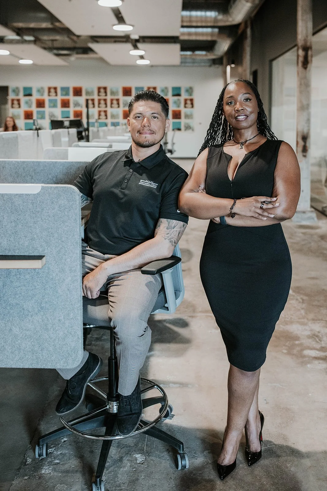 A man and a woman in professional attire posing in an office environment with cubicles, artwork on the wall, and overhead lighting.