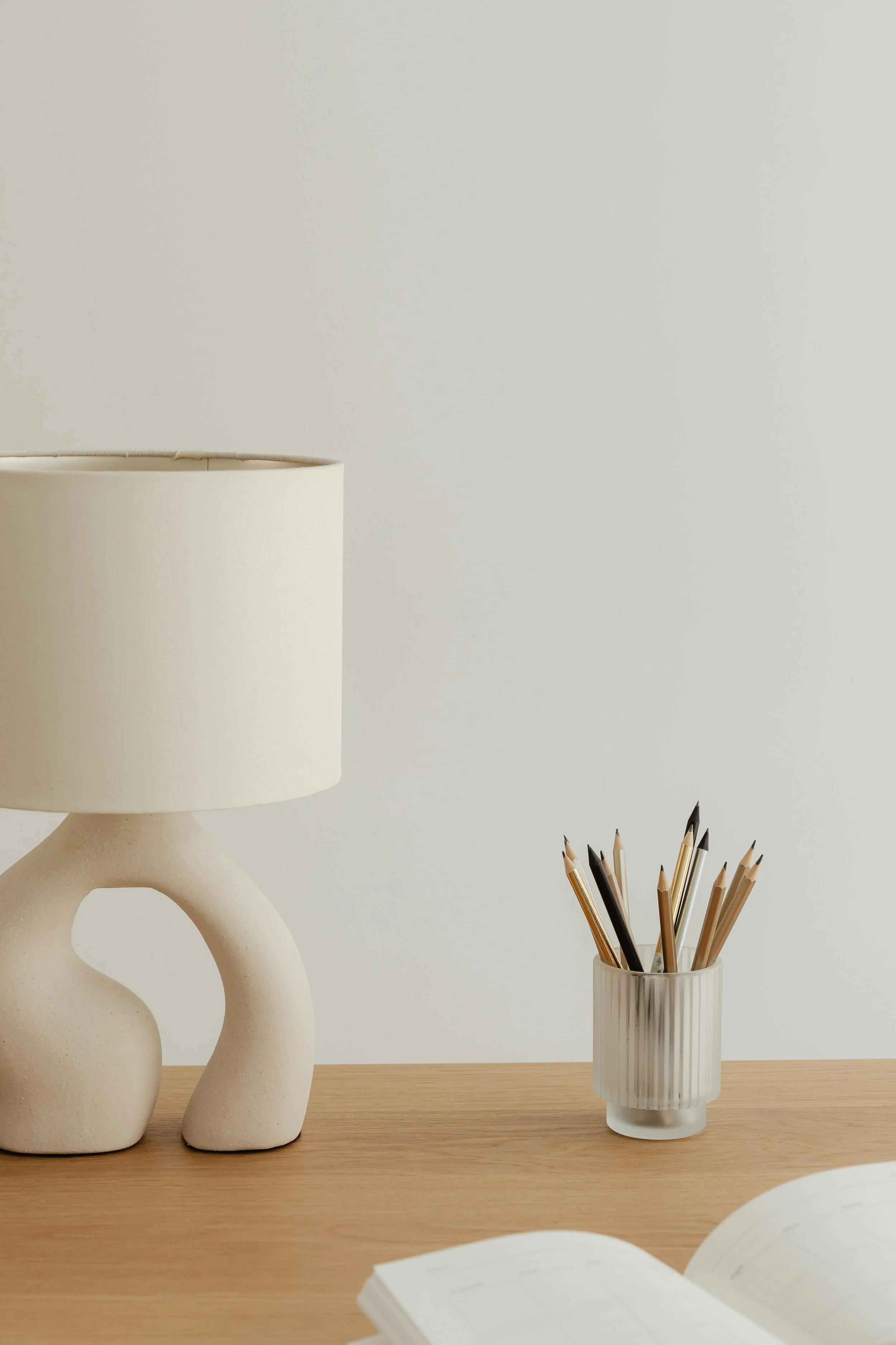 A minimalist wooden desk with a beige lamp and a ribbed white cup filled with pencils, with an open book in the foreground.