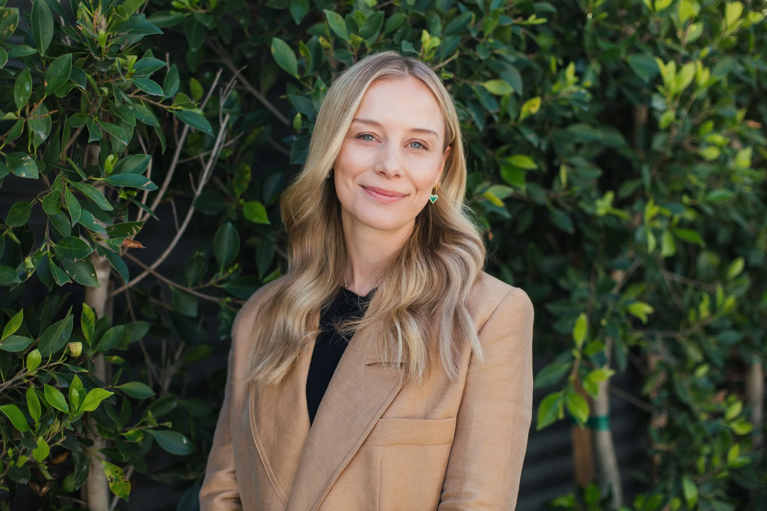 A blonde woman with wavy hair smiling outdoors in front of green leafy bushes, wearing a tan blazer and earrings.
