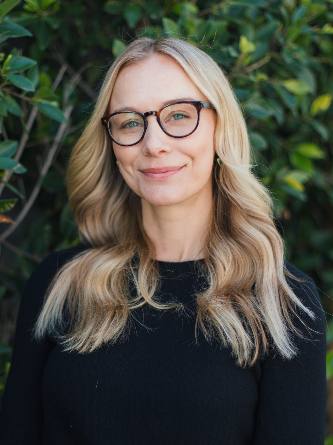 A woman with blonde hair, glasses, and a black top, standing outdoors in front of green foliage, smiling gently.