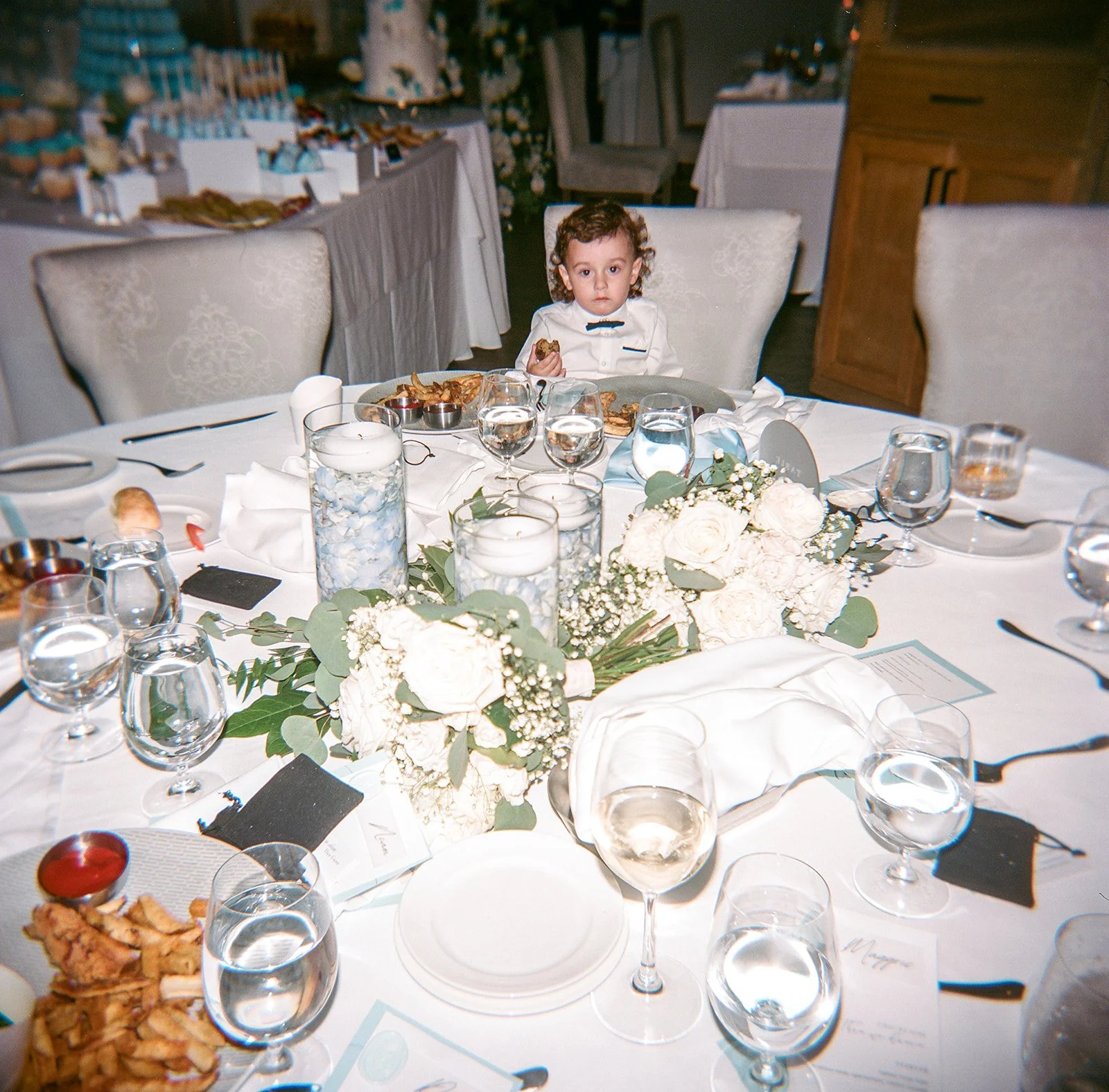 A young boy with curly hair sitting at a decorated banquet table with floral centerpieces, candles, glassware, and plates, holding a piece of food, in an elegant dining room.