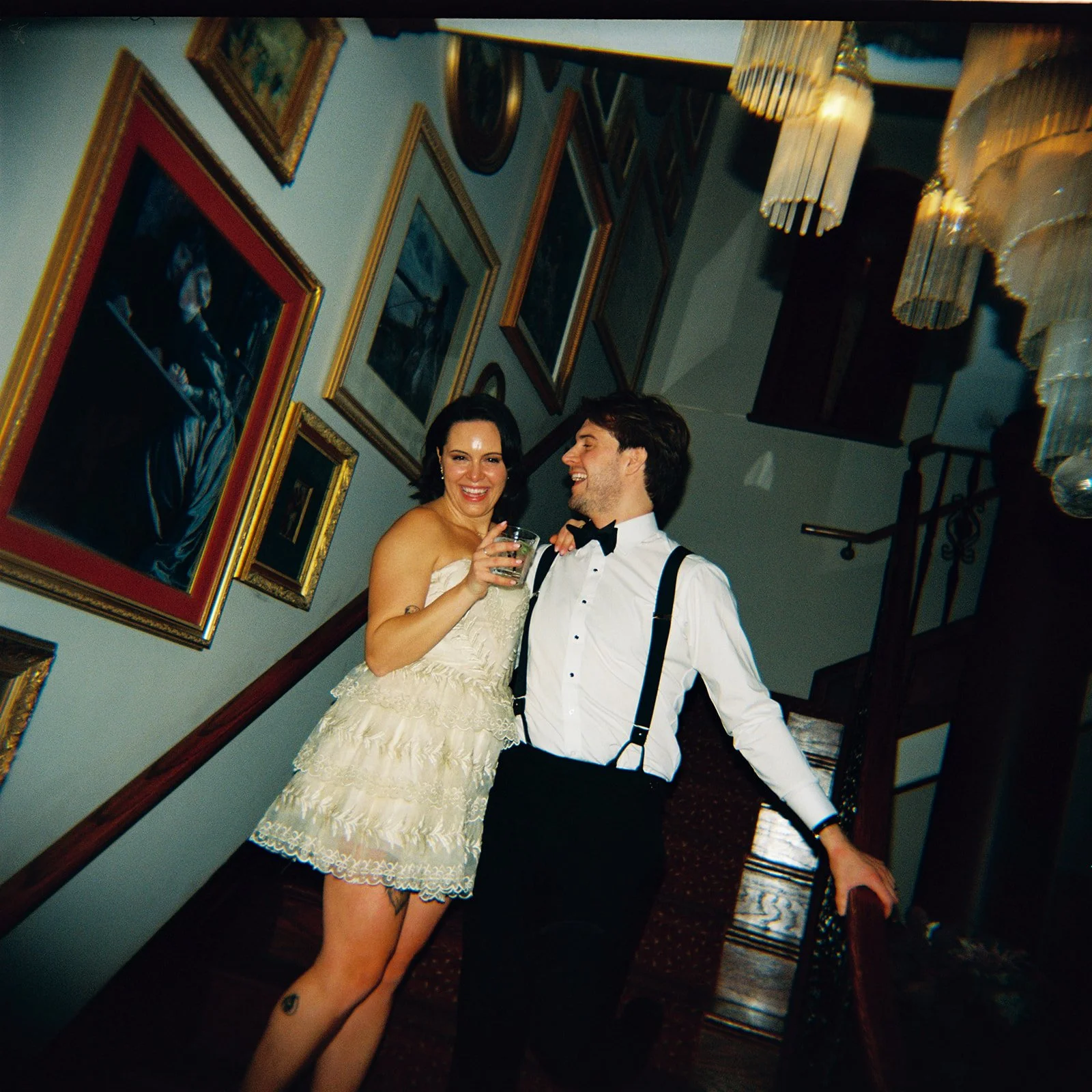 A woman in a cream-colored dress holding a glass, smiling, standing on a staircase next to a man in a white shirt with suspenders and a bow tie, also smiling. They are at a formal gathering or wedding reception, with framed artwork on the wall and ch