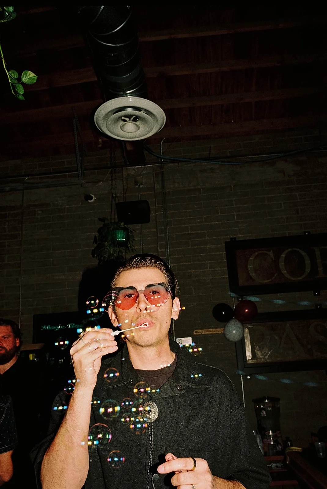 A man wearing sunglasses blowing bubbles at a social gathering or party.
