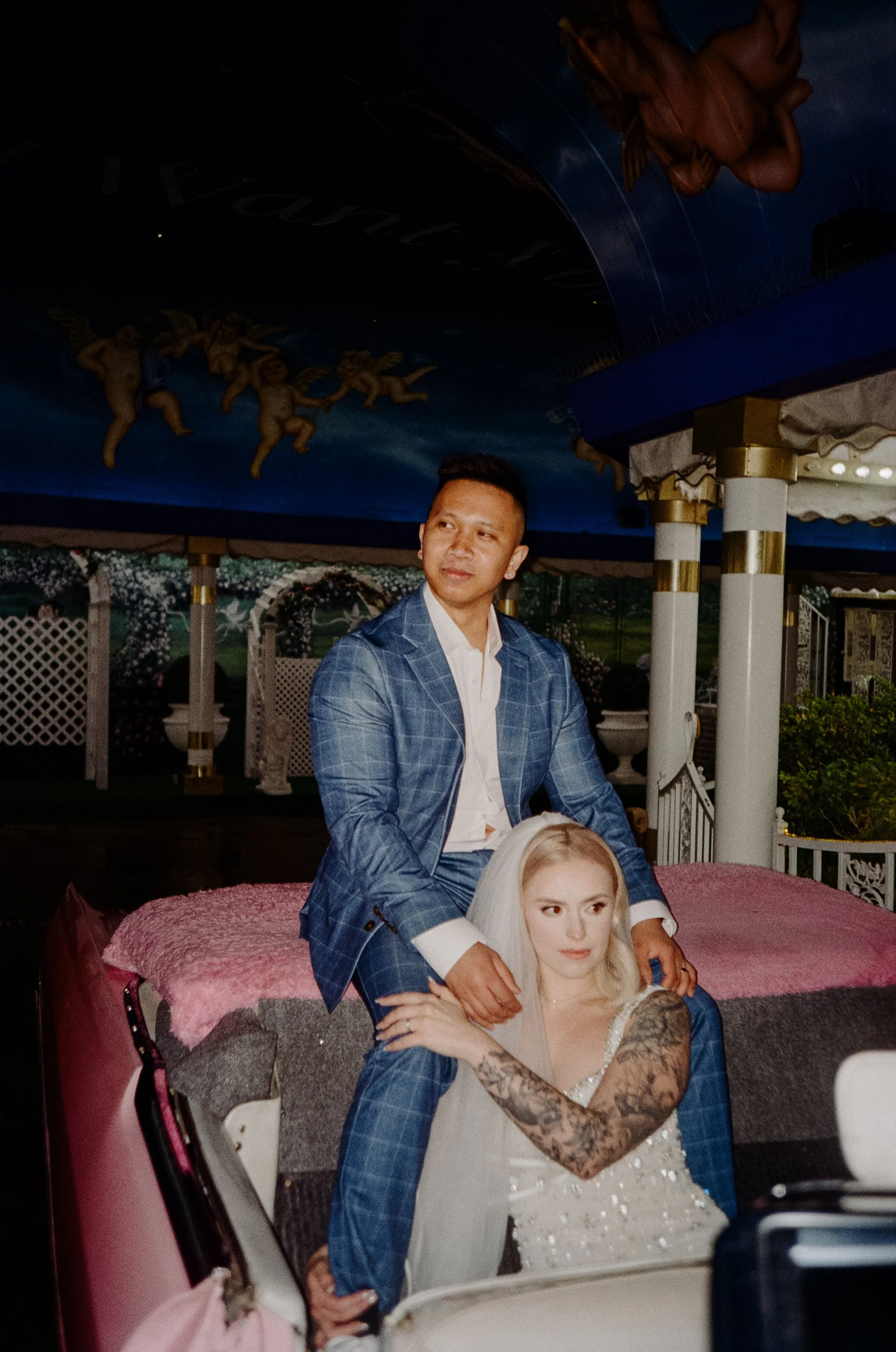 A man in a blue suit sitting on a decorative car with a woman in a white dress and veil, with a painted ceiling featuring cherubs in the background.