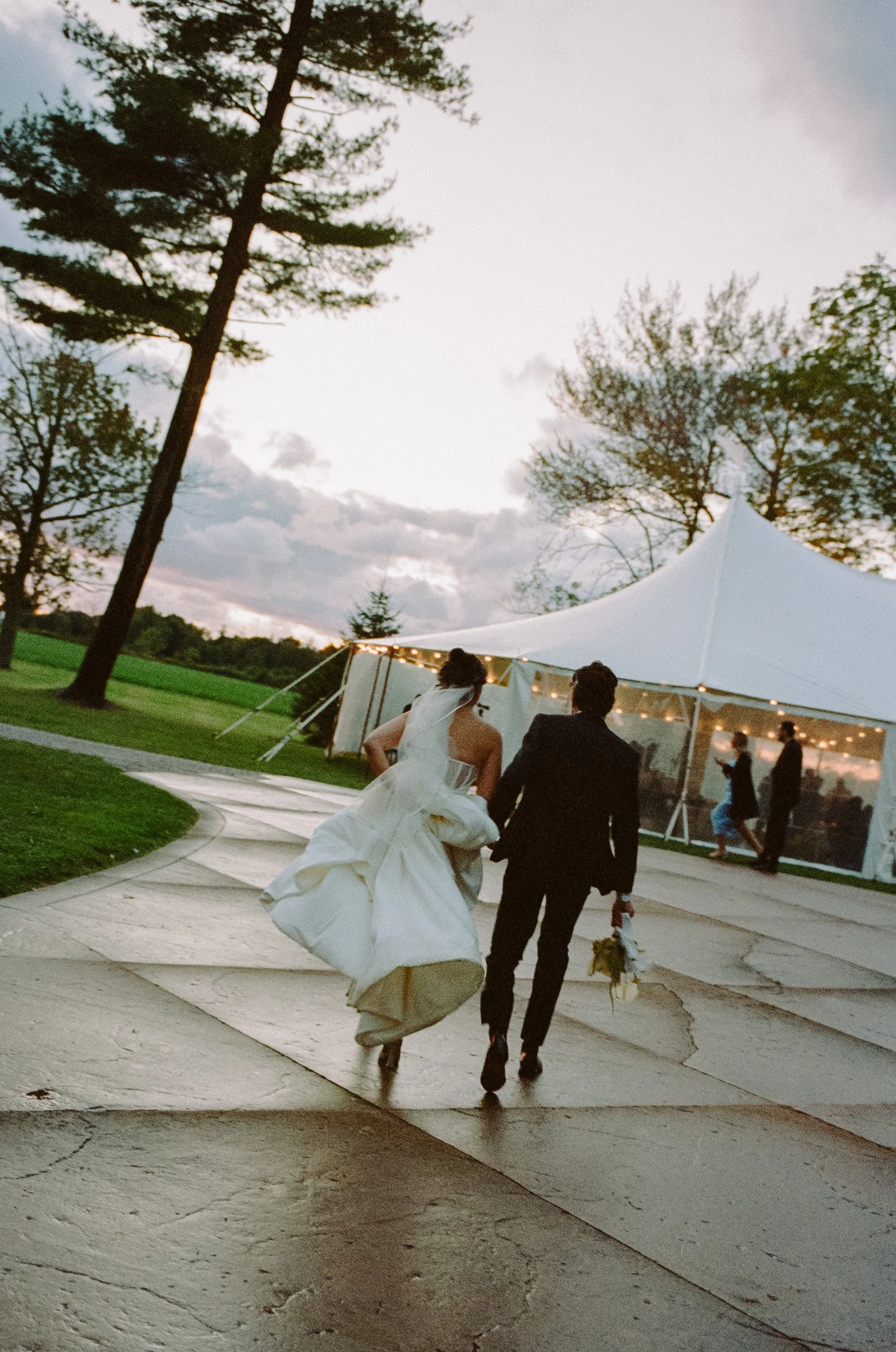 A bride and groom walking away from a wedding reception tent on a winding path, with trees and a cloudy sky in the background.