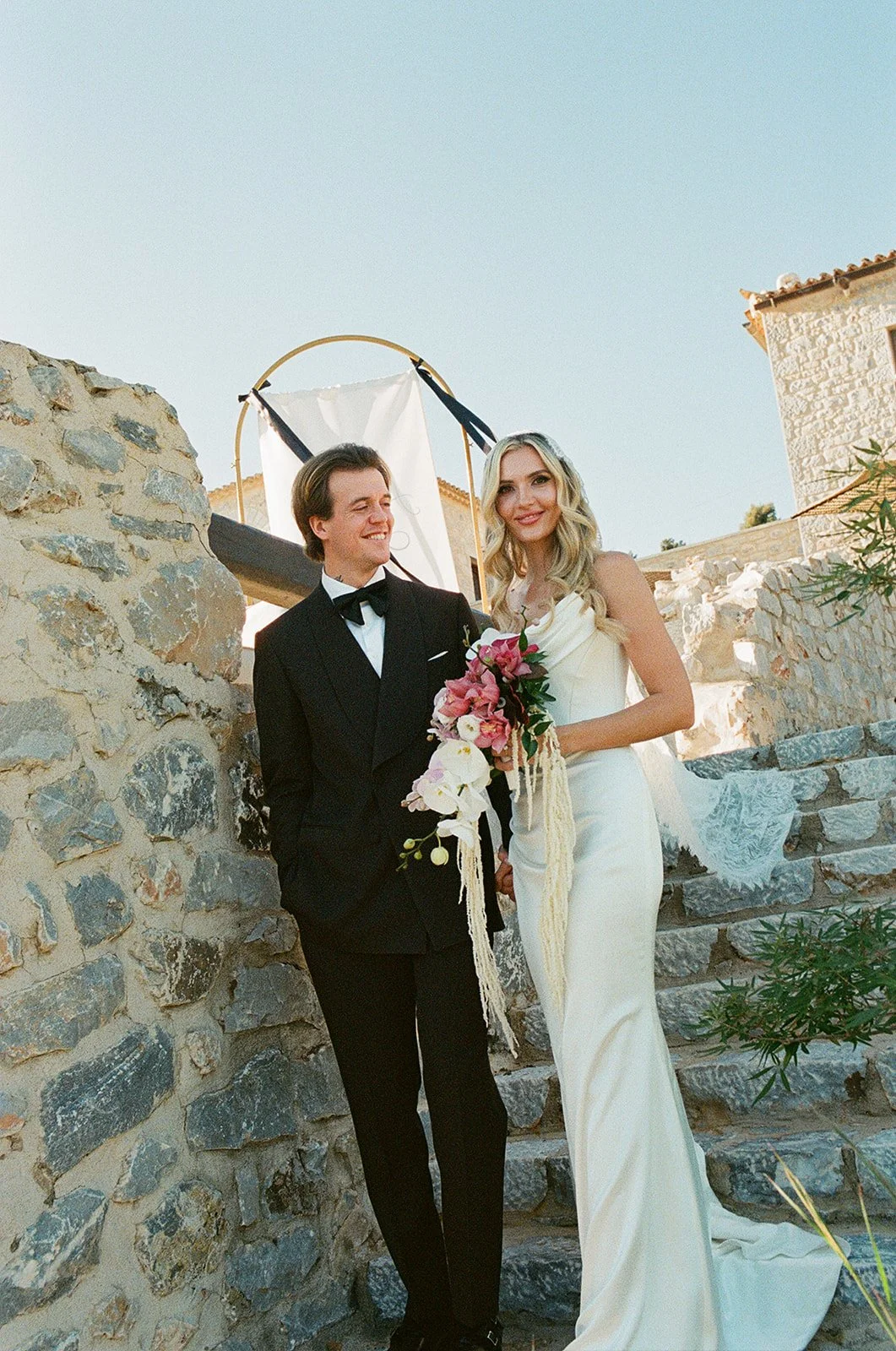 A bride and groom stand together outdoors on stone steps, with the bride holding a bouquet of pink and white flowers, during their wedding celebration.