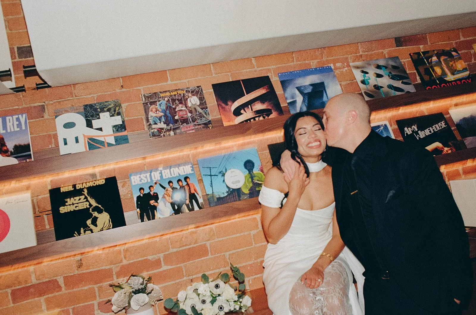A woman in a white dress smiling as a man kisses her cheek in front of a brick wall with vinyl records and album covers displayed on shelves.