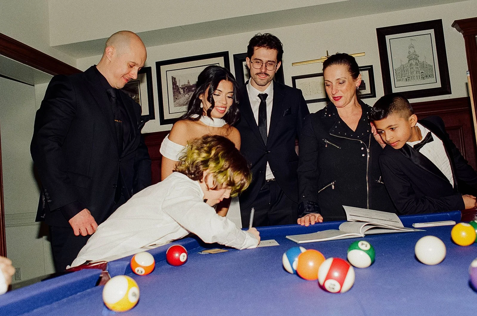 Group of six people gathered around a pool table, watching a young boy in a white shirt take a shot, with pool balls scattered on the table, in a room with framed pictures on the wall.
