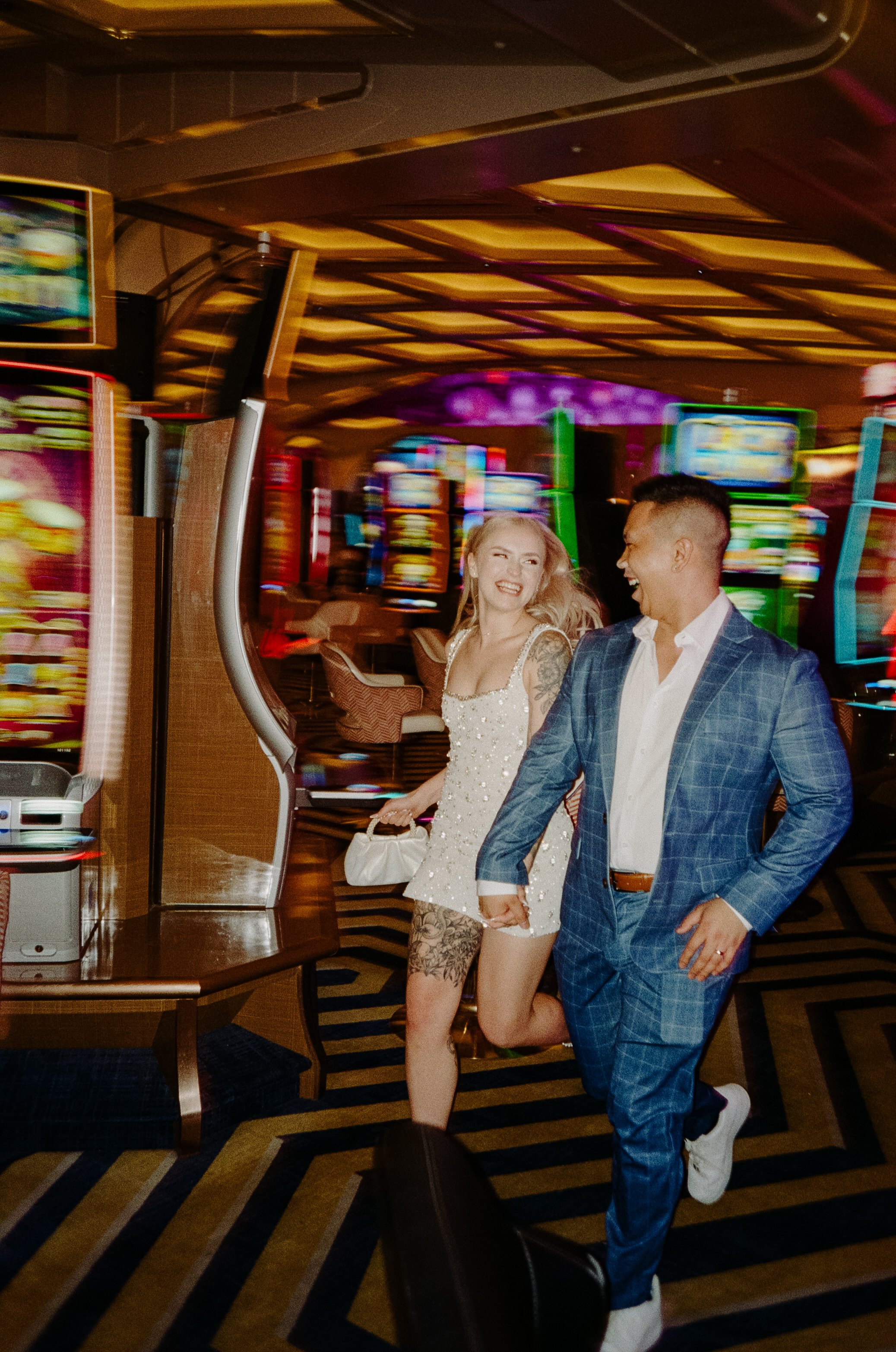 Two happy people, a woman in a white dress and a man in a blue plaid suit, holding hands and running in a brightly lit casino with slot machines and colorful lights.