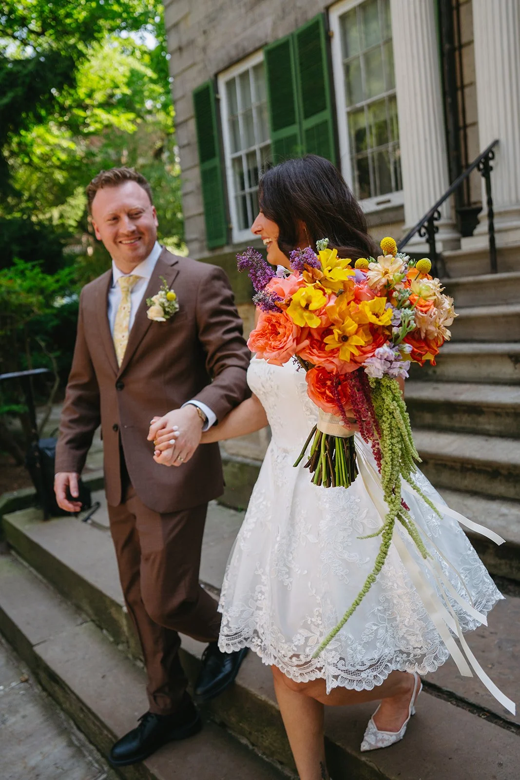 A bride and groom holding hands and walking down steps outside a house, smiling at each other. The bride is carrying a large, colorful bouquet of flowers, and the groom is wearing a suit with a boutonnière.