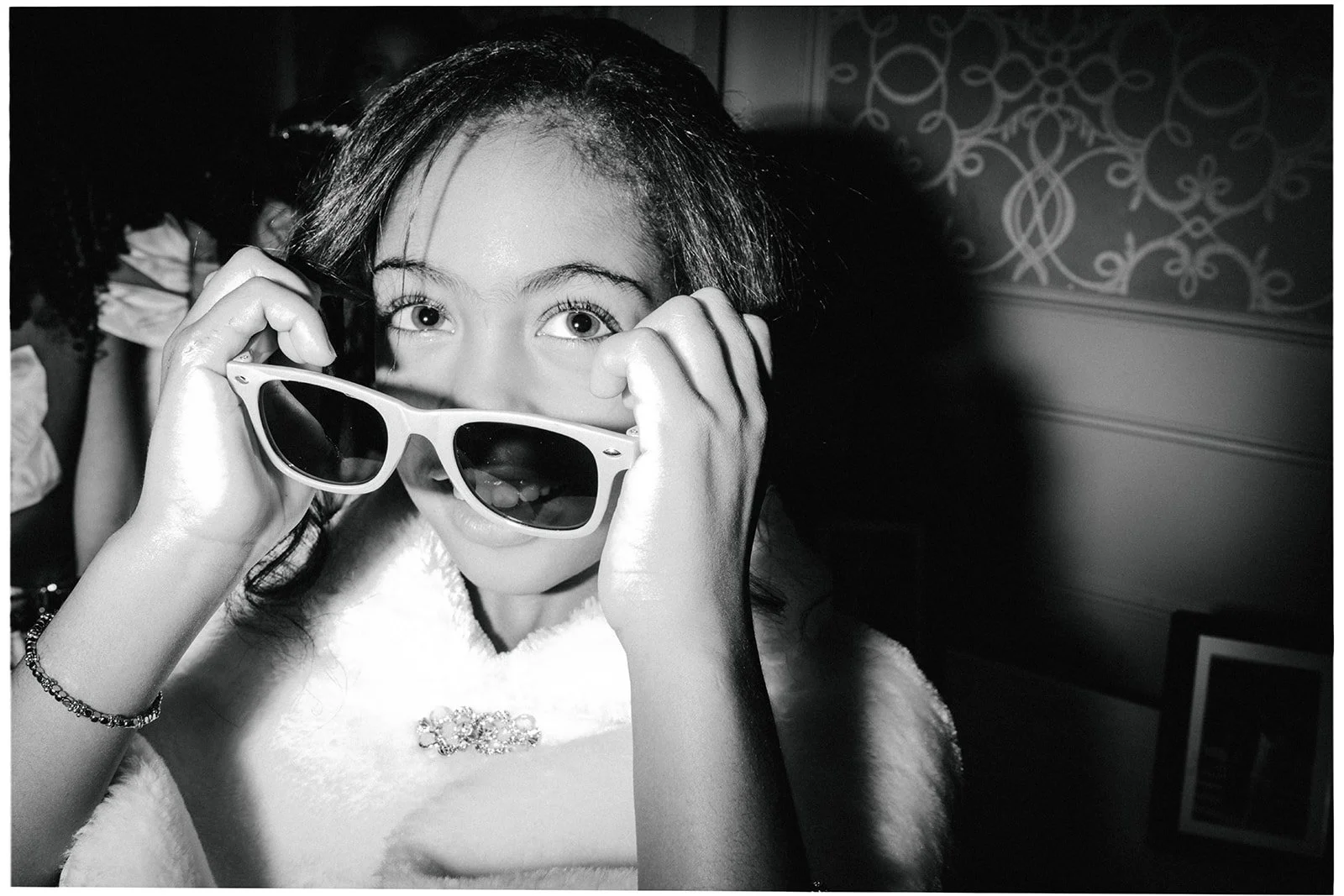 A woman holds a pair of sunglasses in front of her face, looking directly at the camera, in black and white.