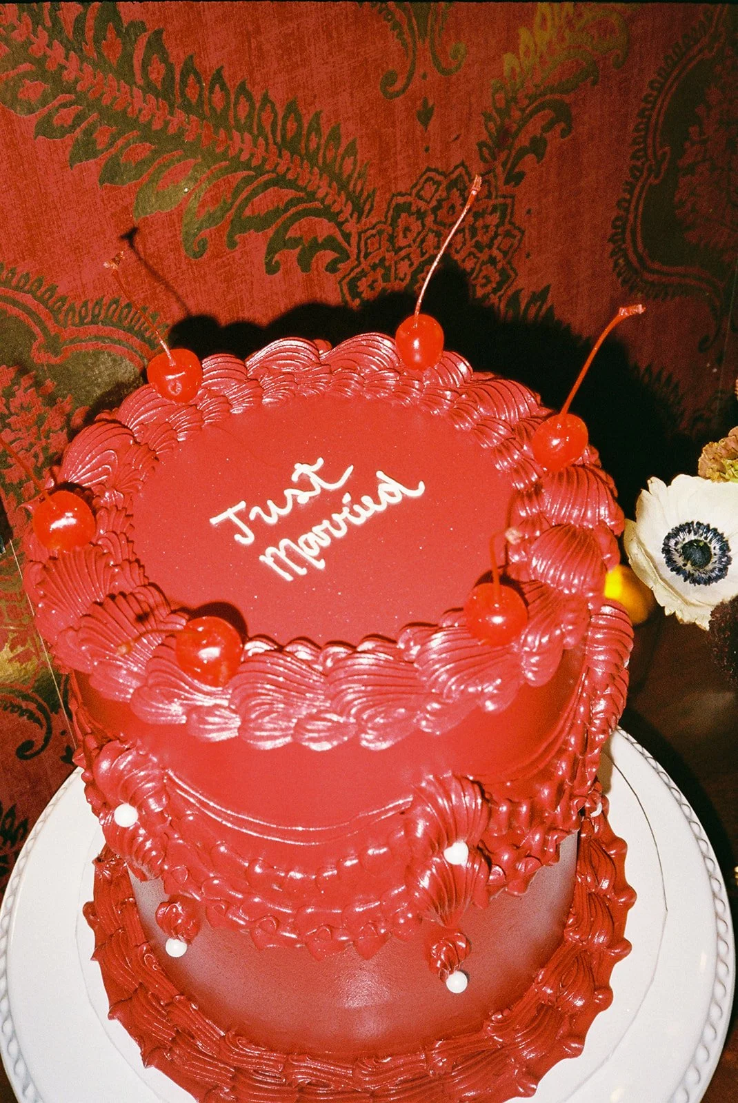 A two-tier red birthday cake with cherry decorations on top. The cake has white writing that says 'Juat Mariano' on the top. It is placed on a white platter against a red patterned background.