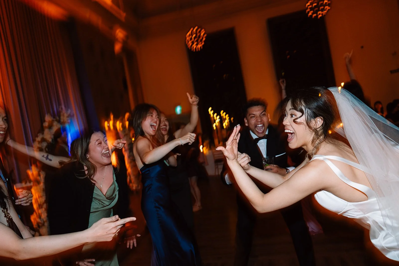 A bride and groom dancing and laughing with friends at a wedding reception in a warmly lit room.