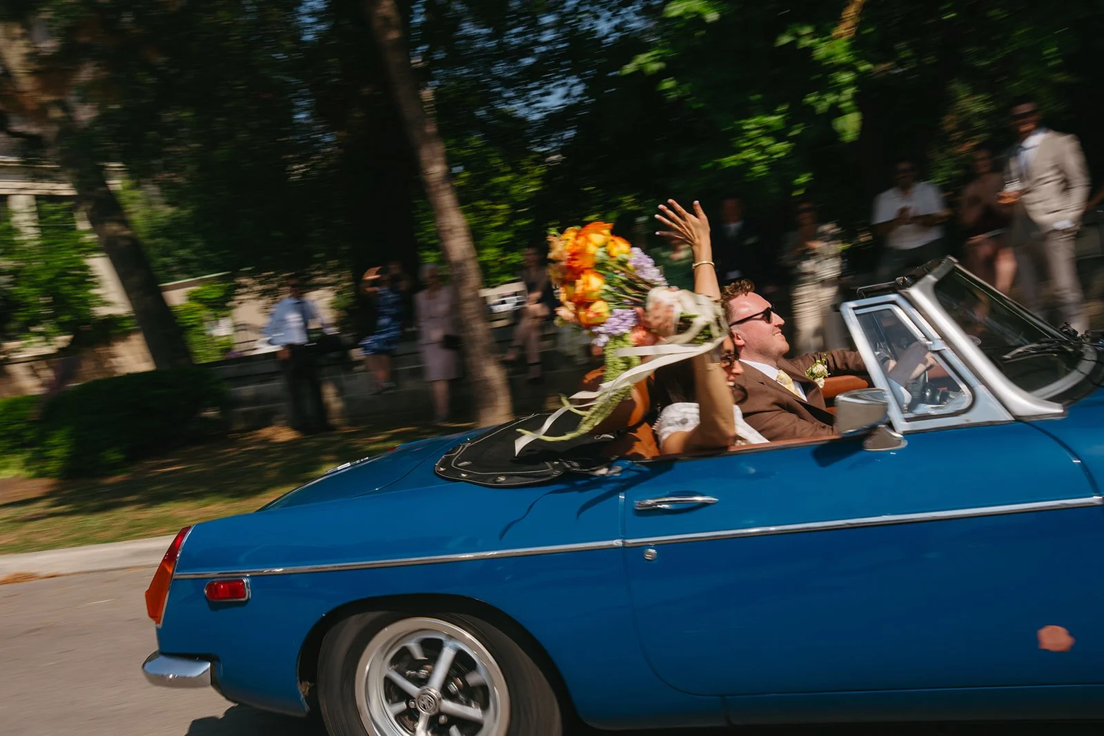 Couple with a large bouquet of flowers in a vintage blue convertible car, waving to onlookers during a parade or celebration.