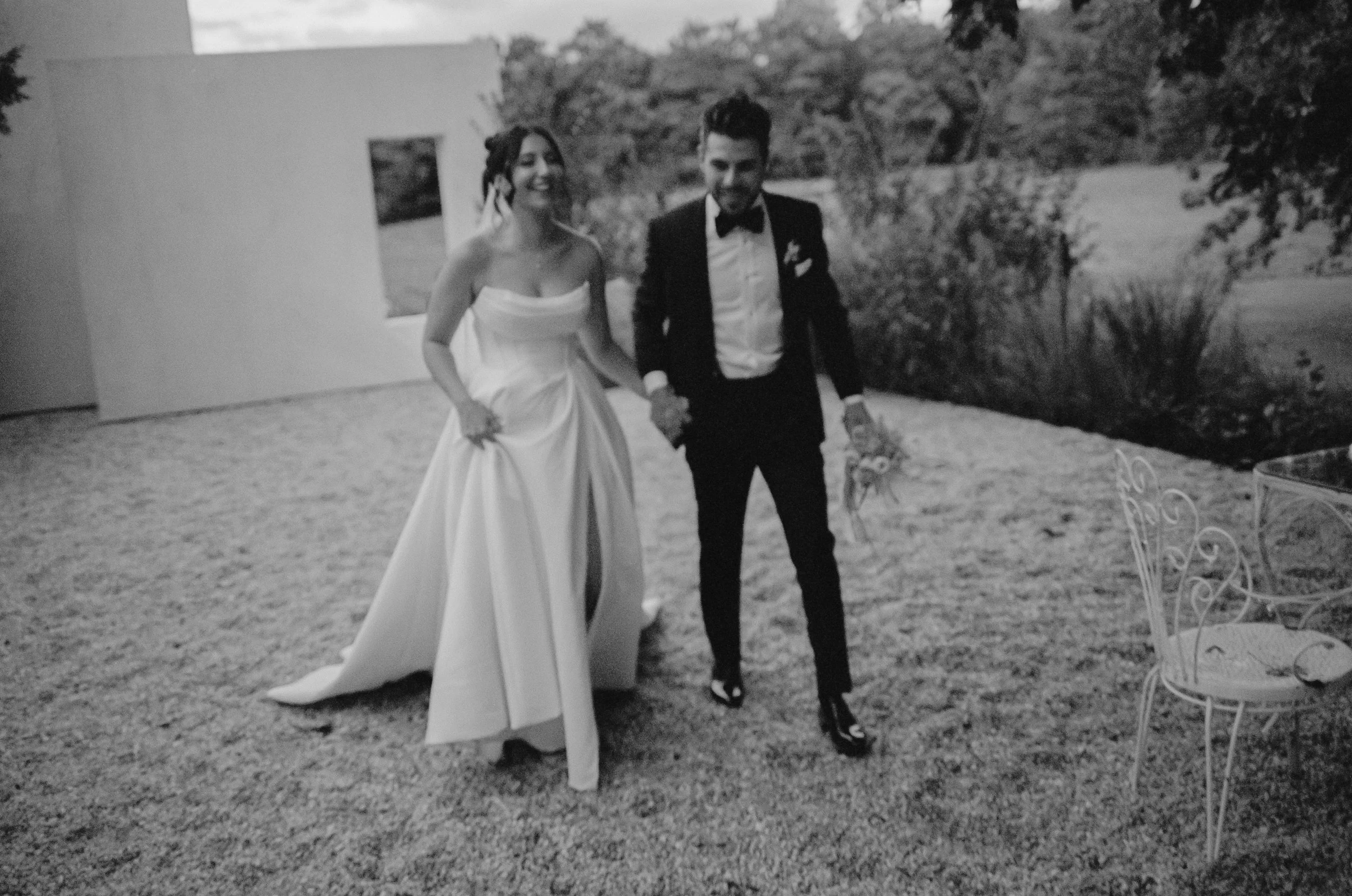 A bride in a strapless wedding dress and a groom in a tuxedo walk hand-in-hand outdoors on grass, smiling, with a backdrop of trees and a building.