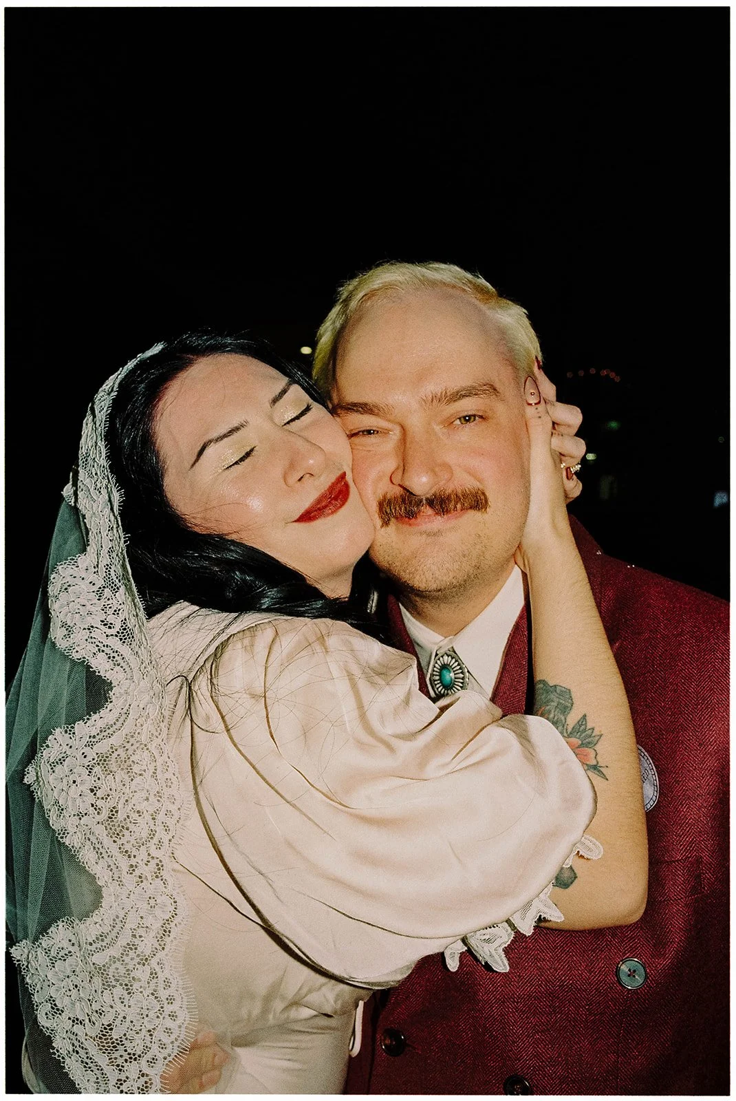 A woman with dark hair wearing a lace veil and a bride's dress is hugging a man with blonde hair and a mustache wearing a maroon blazer. They are close together, with the woman smiling with eyes closed and the man smiling at the camera, against a dar