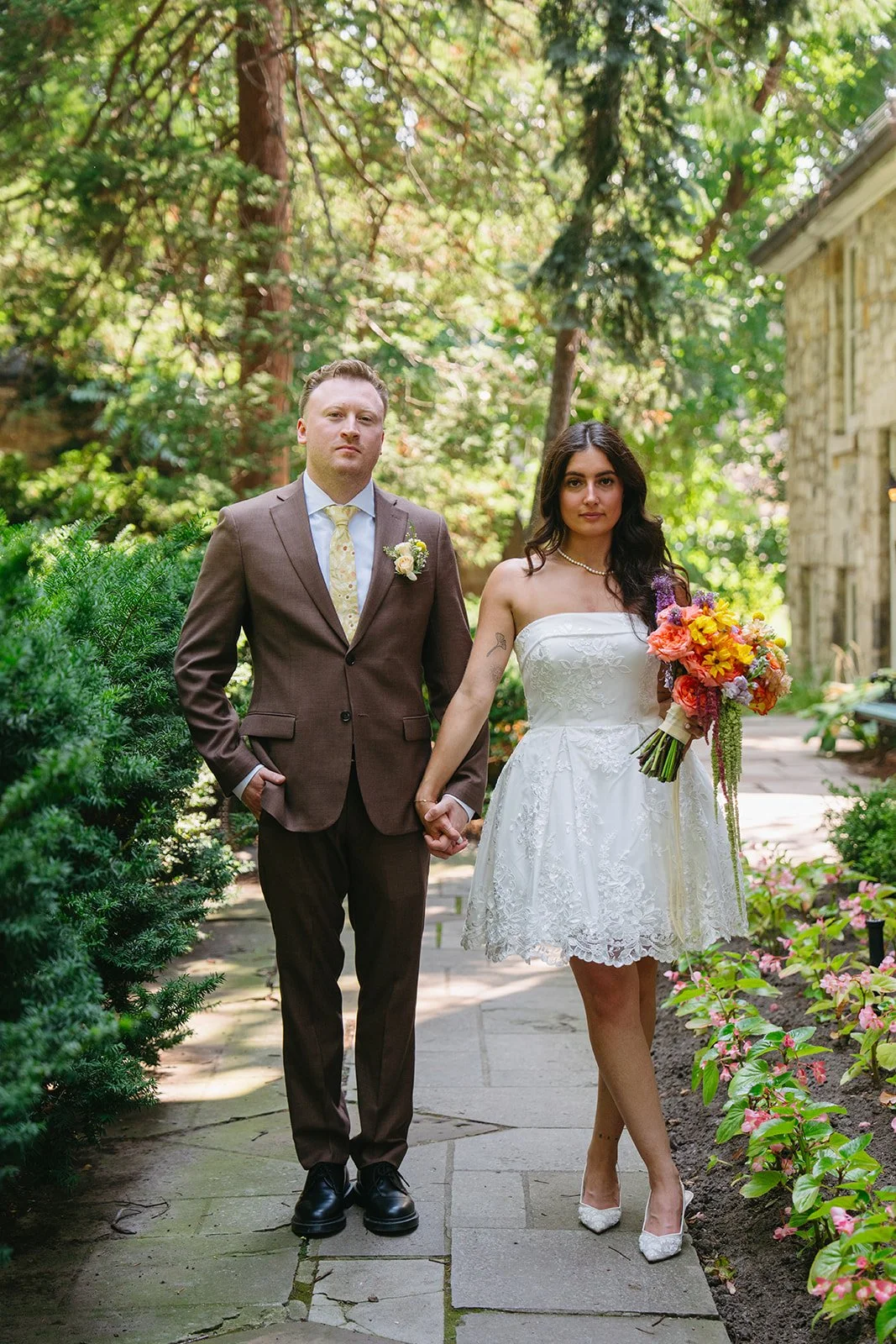 A newlywed couple holding hands on a garden path, surrounded by trees and flowers.