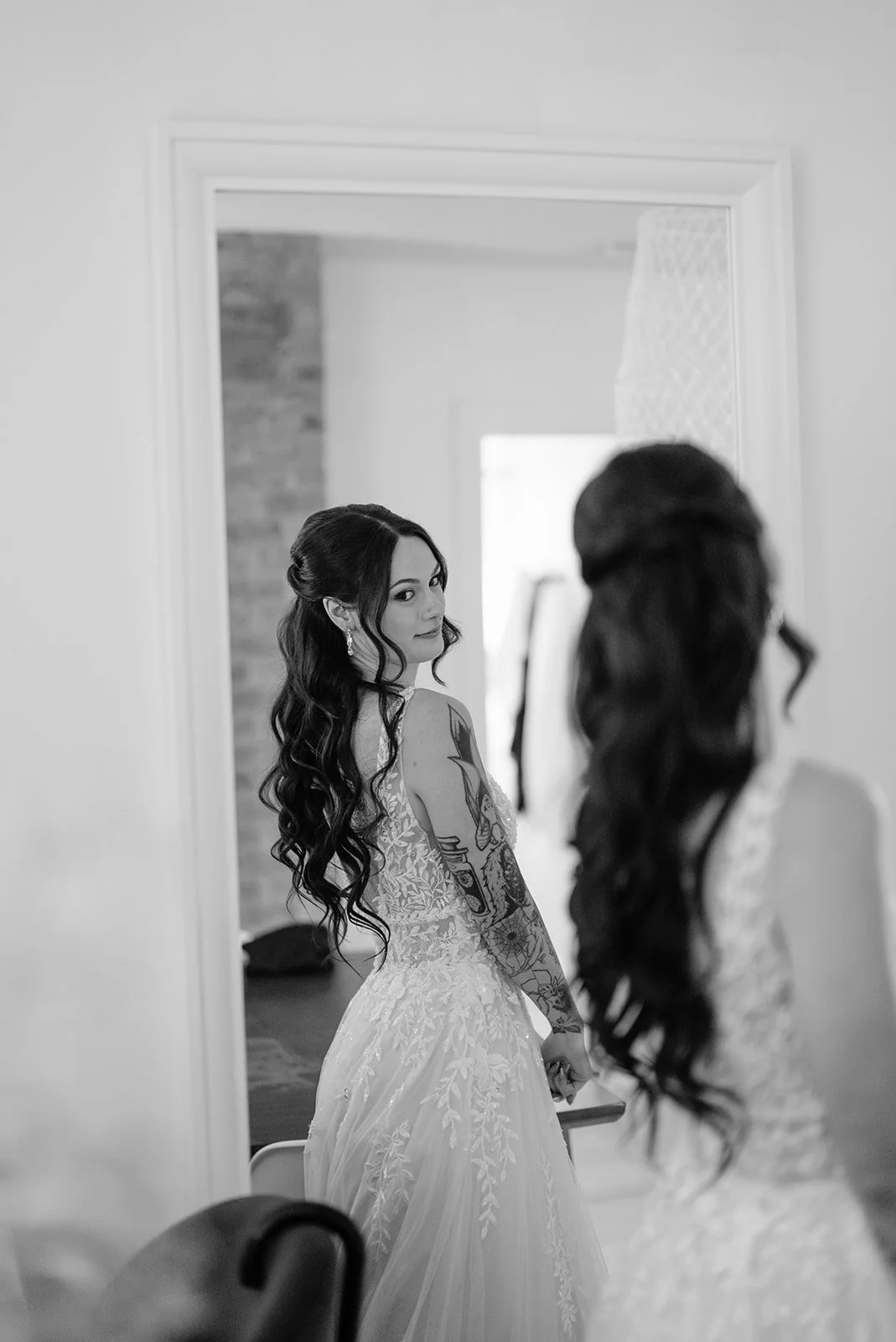 A bride with long dark wavy hair looks into a mirror while getting ready for her wedding, wearing a lace wedding gown with tattoos on her arm.