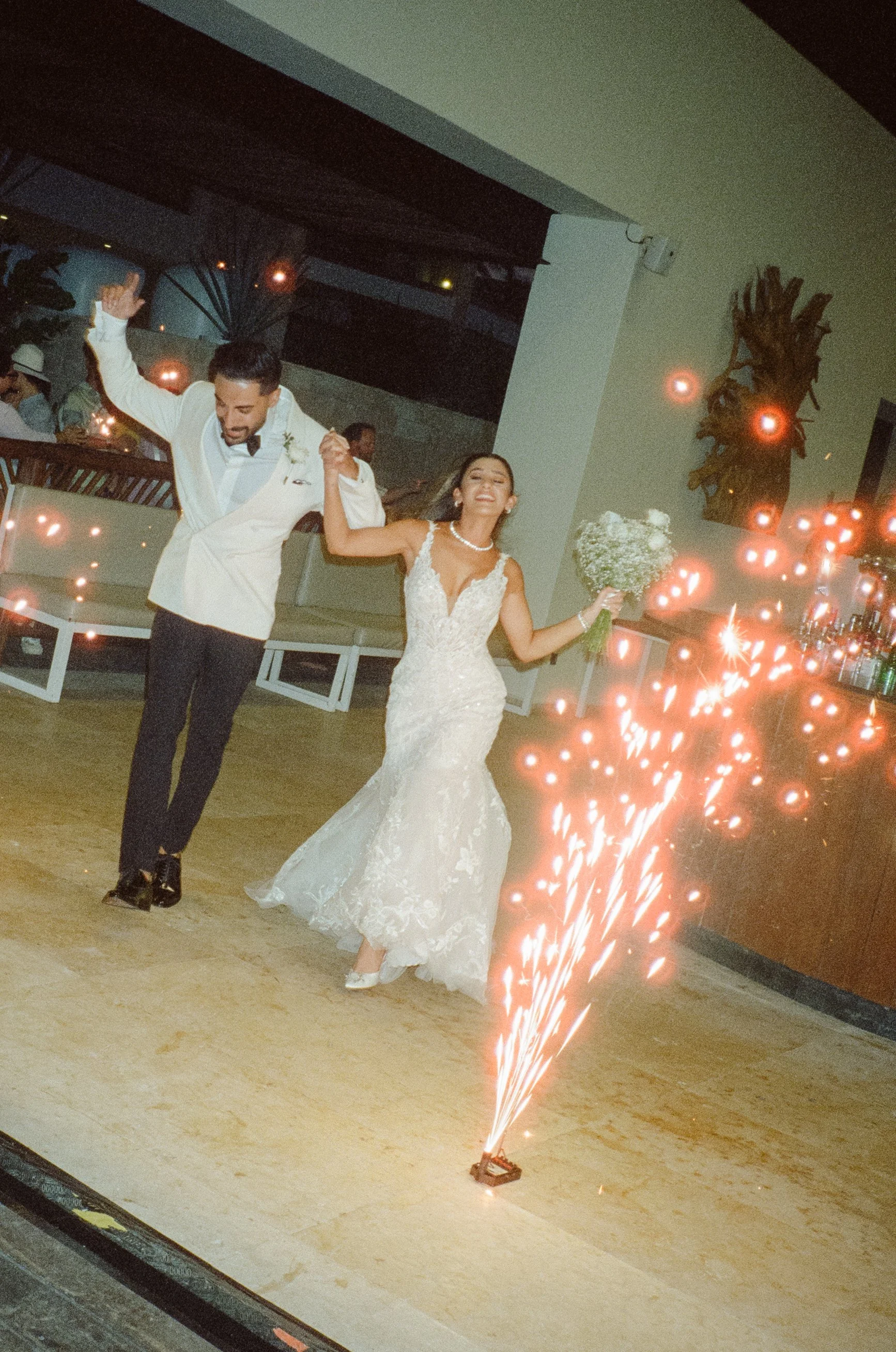 A bride and groom dancing at their wedding reception, holding hands and smiling, with sparklers on the floor in front of them.