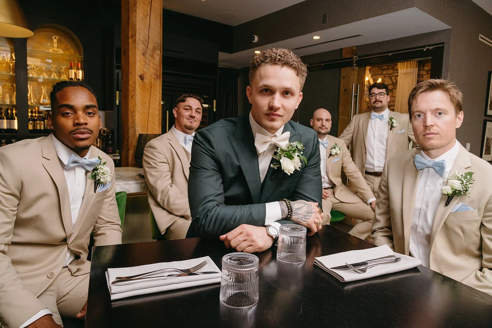A group of seven men dressed in formal suits, some wearing cream-colored tuxedos with light blue bow ties and one in a dark suit with a white bow tie, sitting and standing around a dark wooden table in a dimly lit room with warm decor, including wood