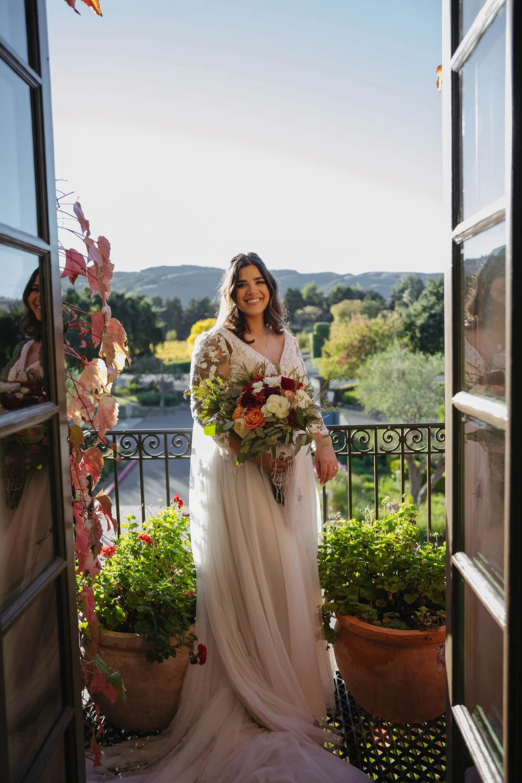 A smiling bride in a white wedding dress holding a bouquet of flowers on a balcony with a scenic outdoor view.