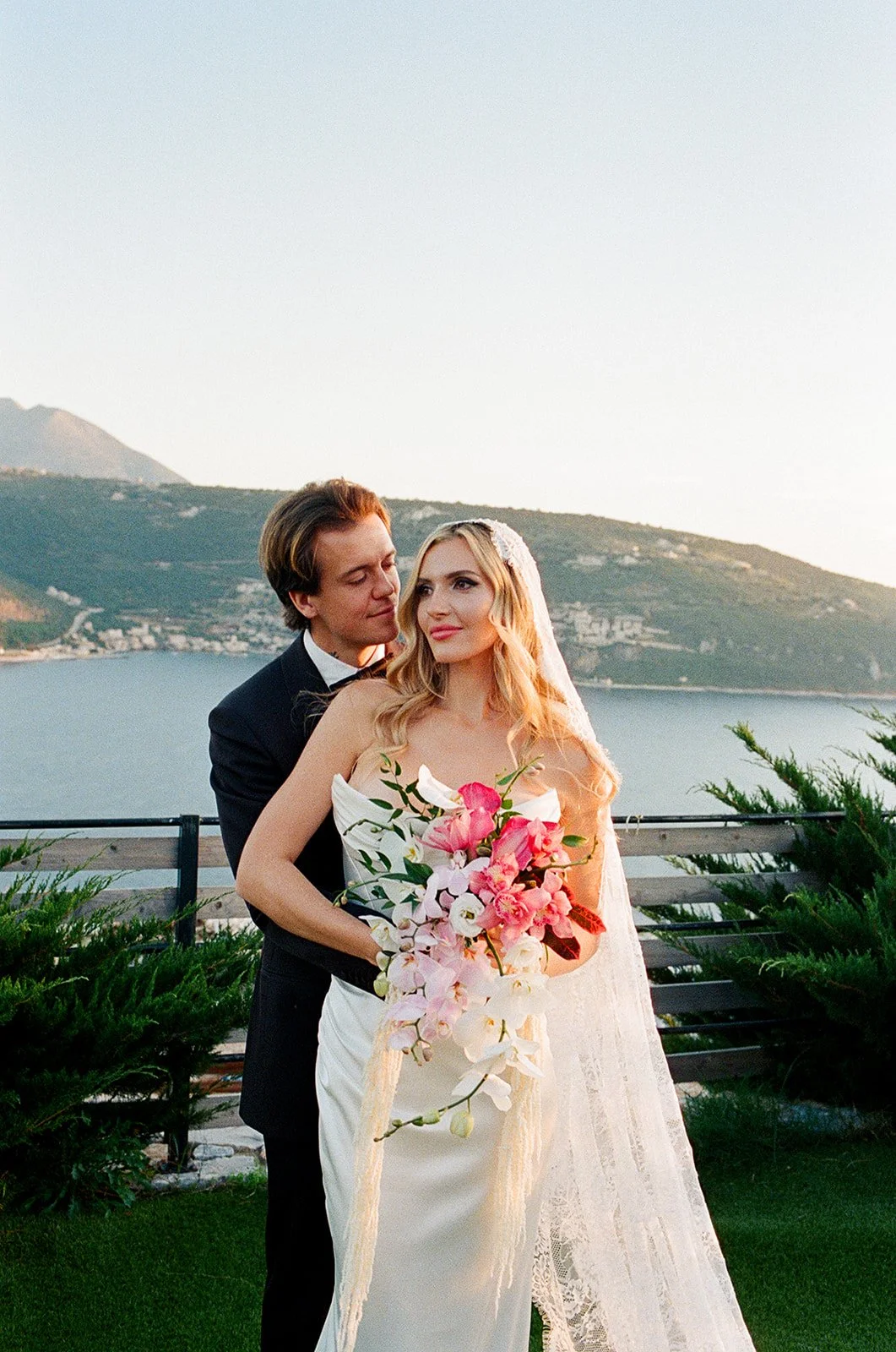 A bride and groom standing outdoors by a lake, with green bushes and mountains in the background. The bride holds a bouquet of pink and white flowers, and both are dressed in wedding attire.