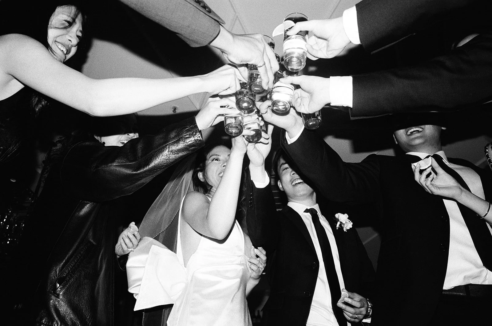 Black and white photo of a group of people celebrating, raising glasses in a toast, smiling and laughing, at a wedding or party.